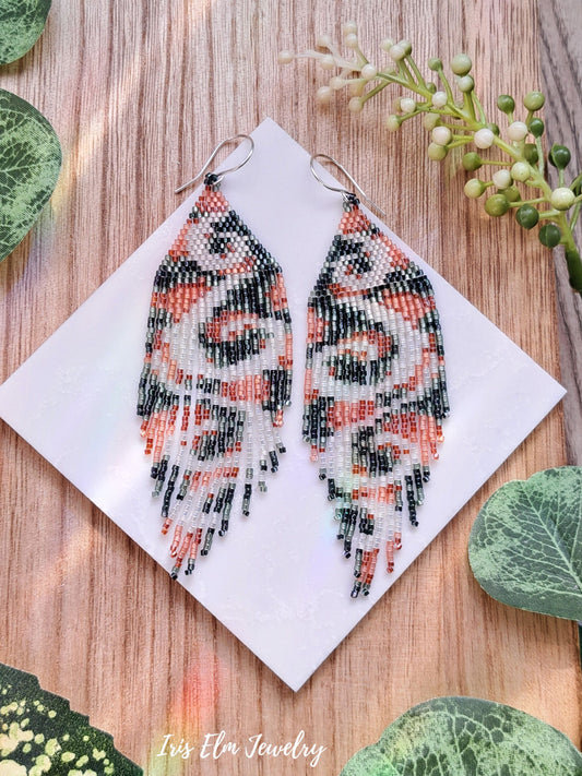 Beaded peach green white swirl long seed bead fringe earrings on a white card with green leaves around on a wooden surface
