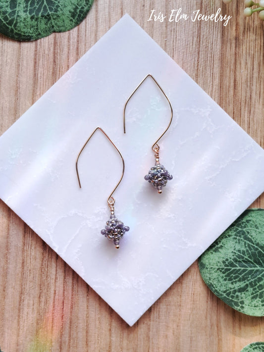 Pair of gold earrings with purple glass beads on a white tile with 'Iris Elm Jewelry' branding.