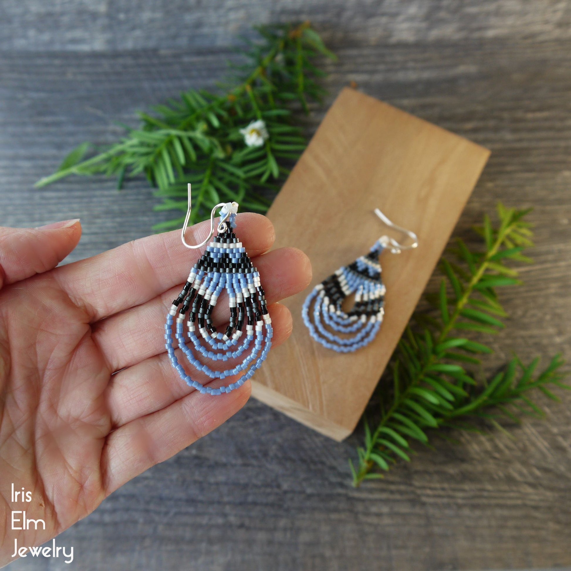 light blue and black Beaded teardrop earrings held by a hand with a wooden block and greenery in the background, featuring 'Iris Elm Jewelry' branding.