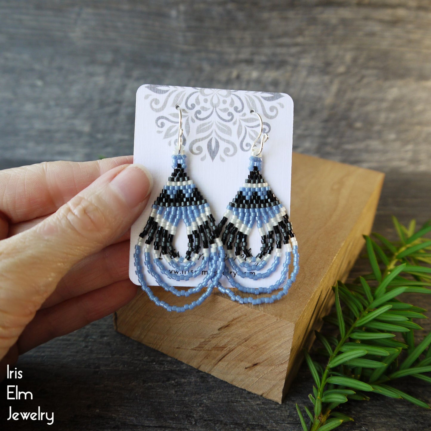 Pair of beaded woven earrings on a card held by a hand with a wooden block and greenery in the background.