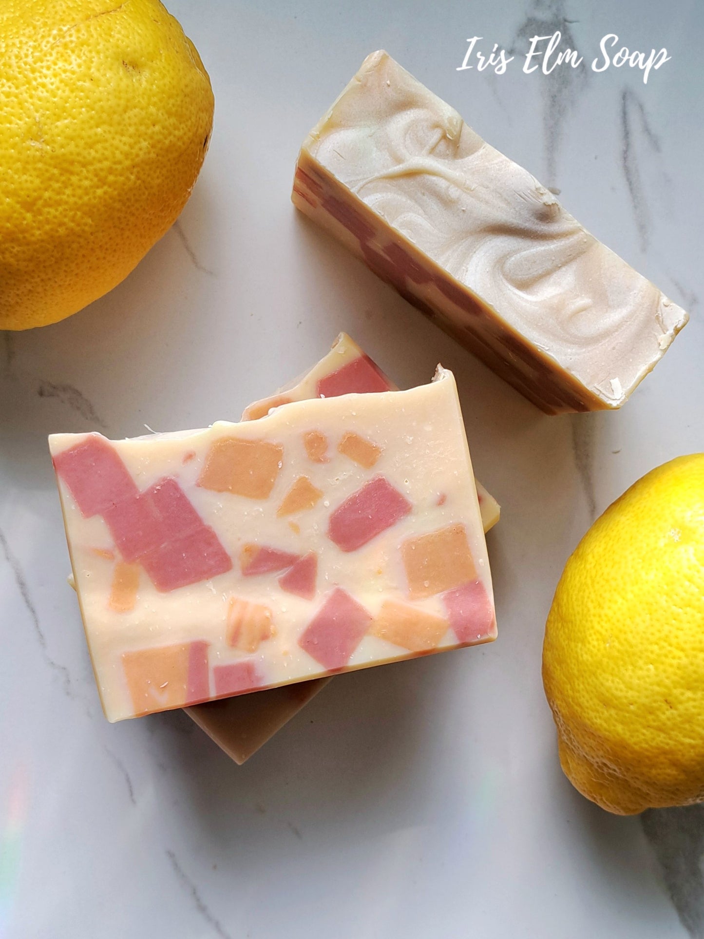 Two bars of sweet lemon tea handmade soap with a colorful pattern next to two lemons on a marble surface, with 'Iris Elm Soap' branding.