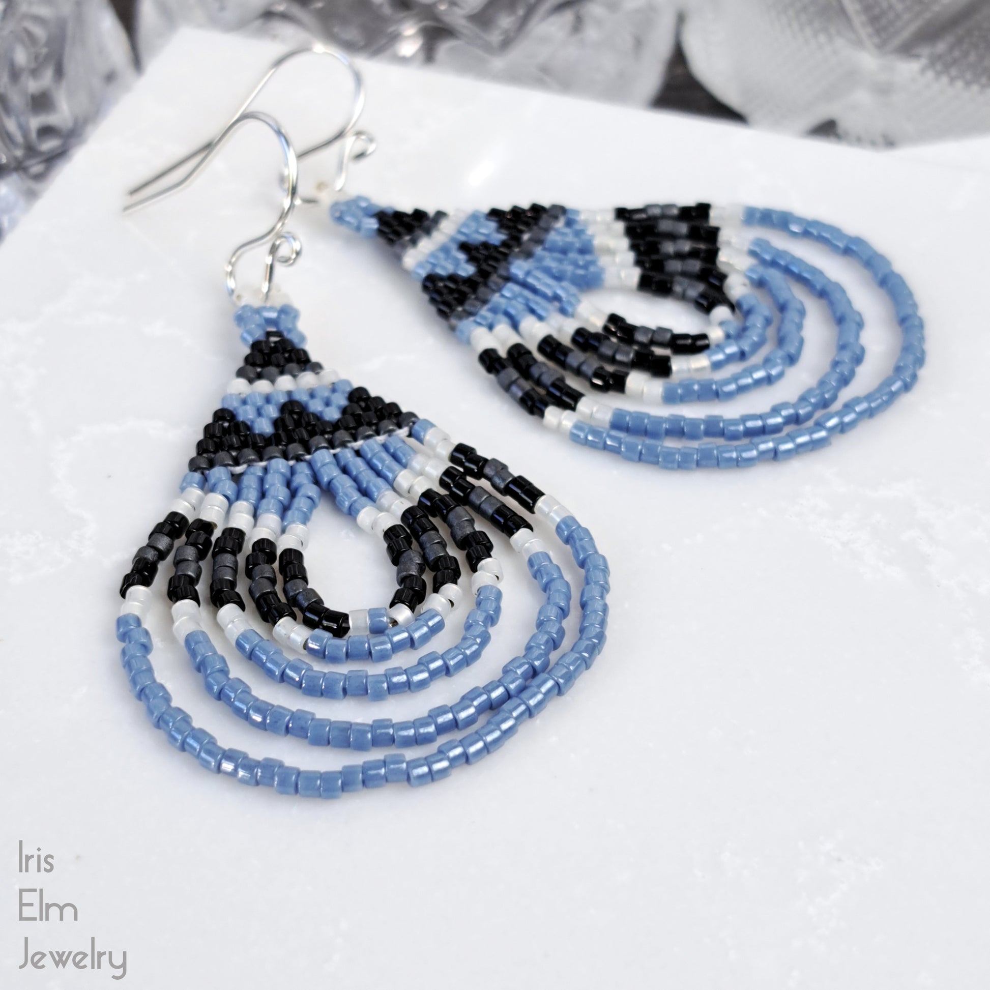 Beaded earrings with blue and black pattern and sterling silver ear wires on a white surface, brand 'Iris Elm Jewelry' visible.