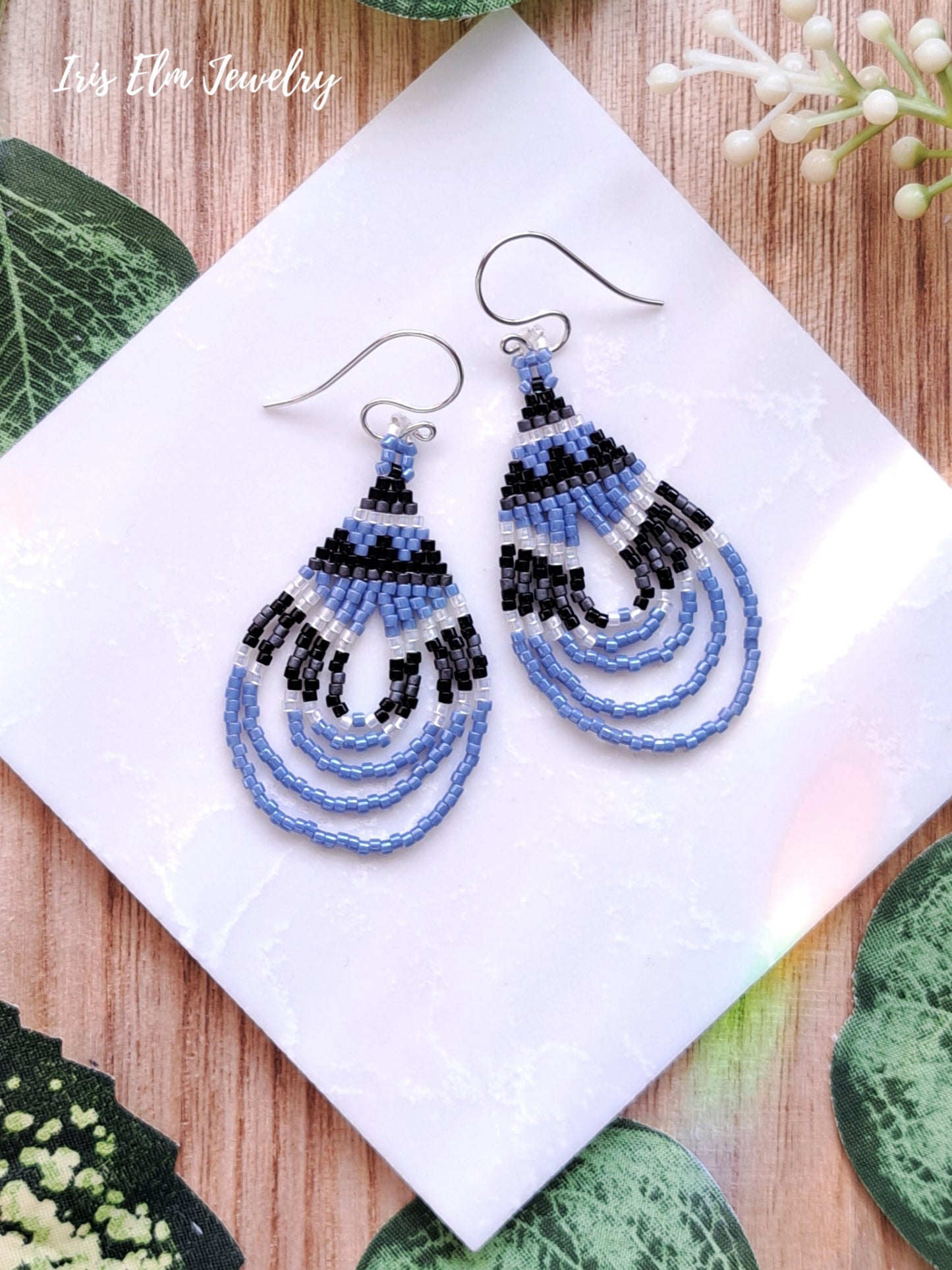 Beaded light blue seed bead woven teardrop earrings on a white tile with green leaves and white flowers in the background