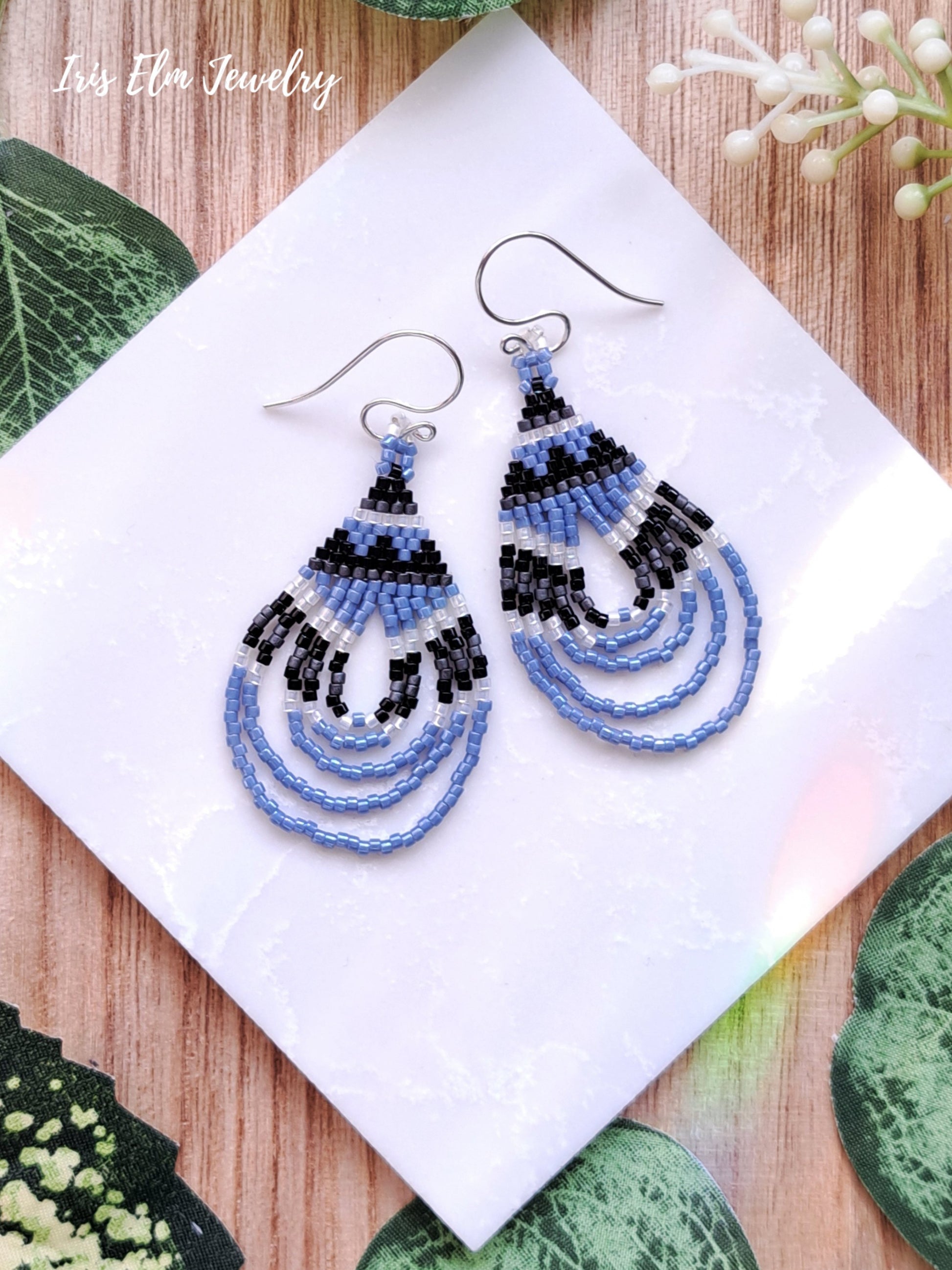 Beaded light blue seed bead woven teardrop earrings on a white tile with green leaves and white flowers in the background
