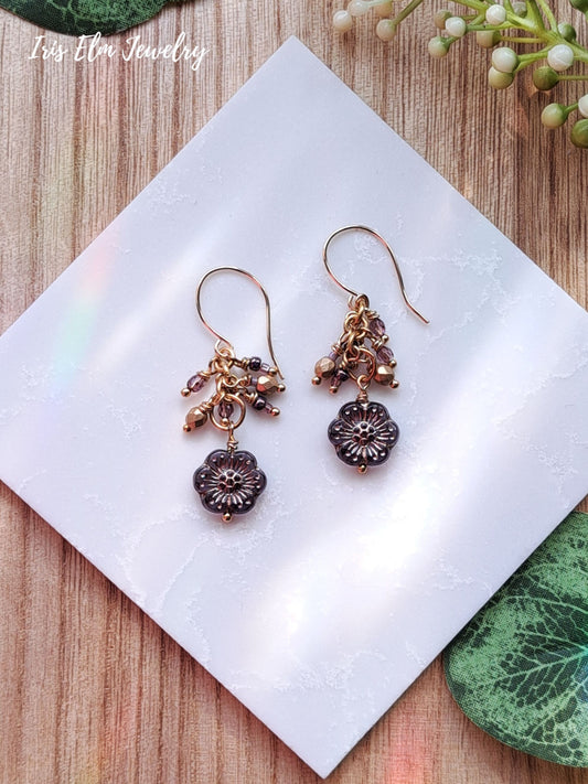 Pair of gold and purple glass cluster floral earrings on a white tile with 'Iris Elm Jewelry' branding.