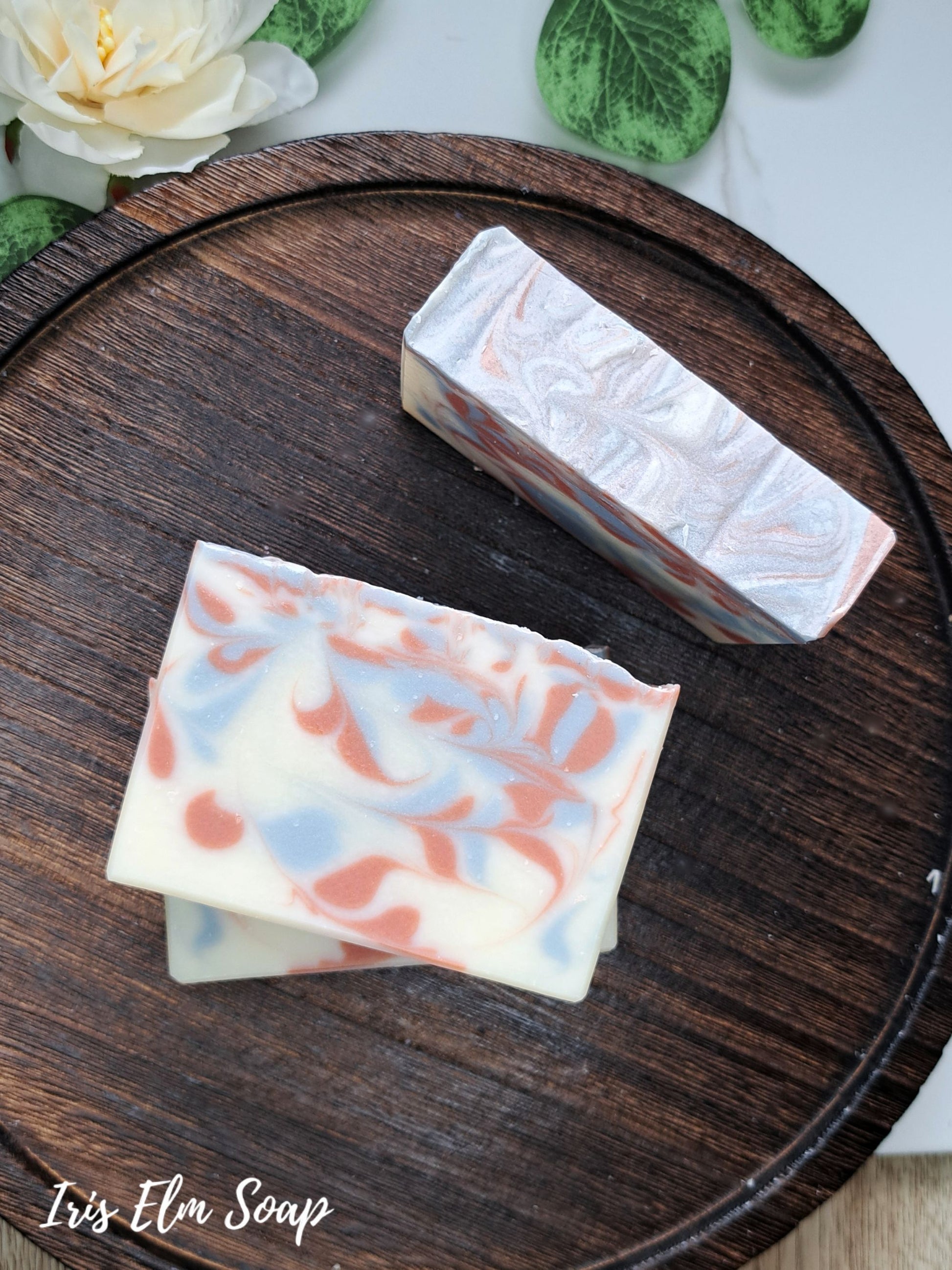 Two rectangular bars of swirled artisan soap on a round dark wooden tray, with a white flower and green leaves in the background. The brand name 'Iris Elm Soap' is visible in the bottom-left corner.