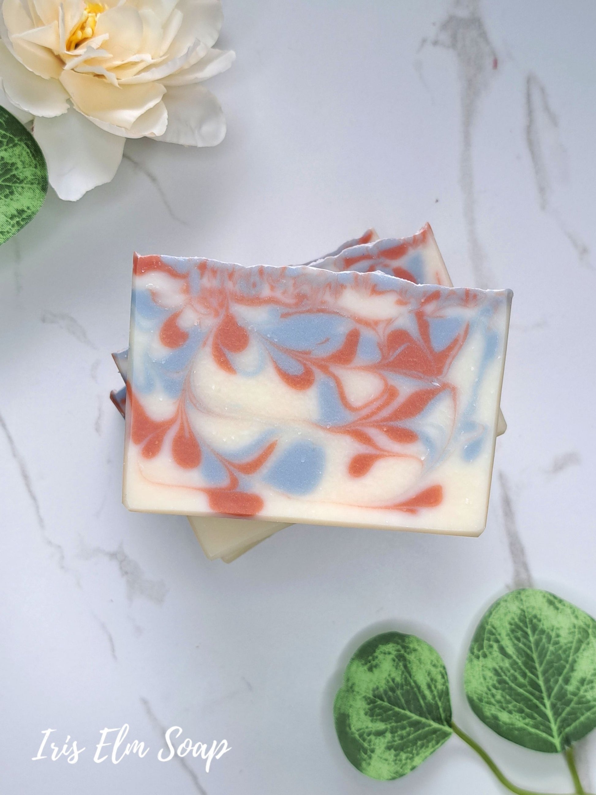 Two rectangular bars of swirled handmade soap with cream, light blue, and coral-red patterns, stacked on a white marble surface with decorative flowers and green leaves. The text 'Iris Elm Soap' appears in the lower left corner.