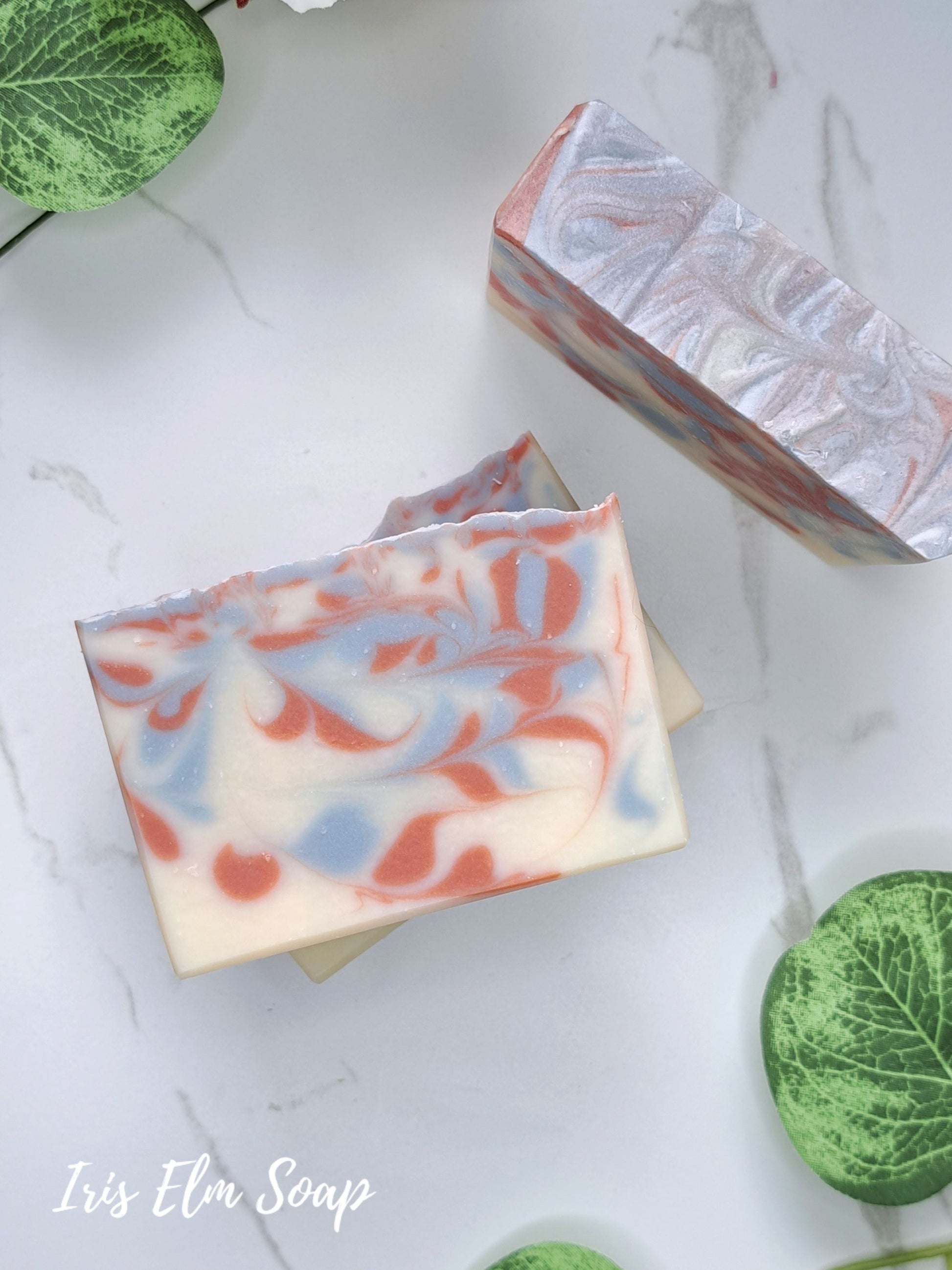 Two handmade soap bars with a marbled swirl pattern of cream, light blue, and coral red, styled on a white marble background with green leaves. The brand name 'Iris Elm Soap' is visible in the bottom left corner.