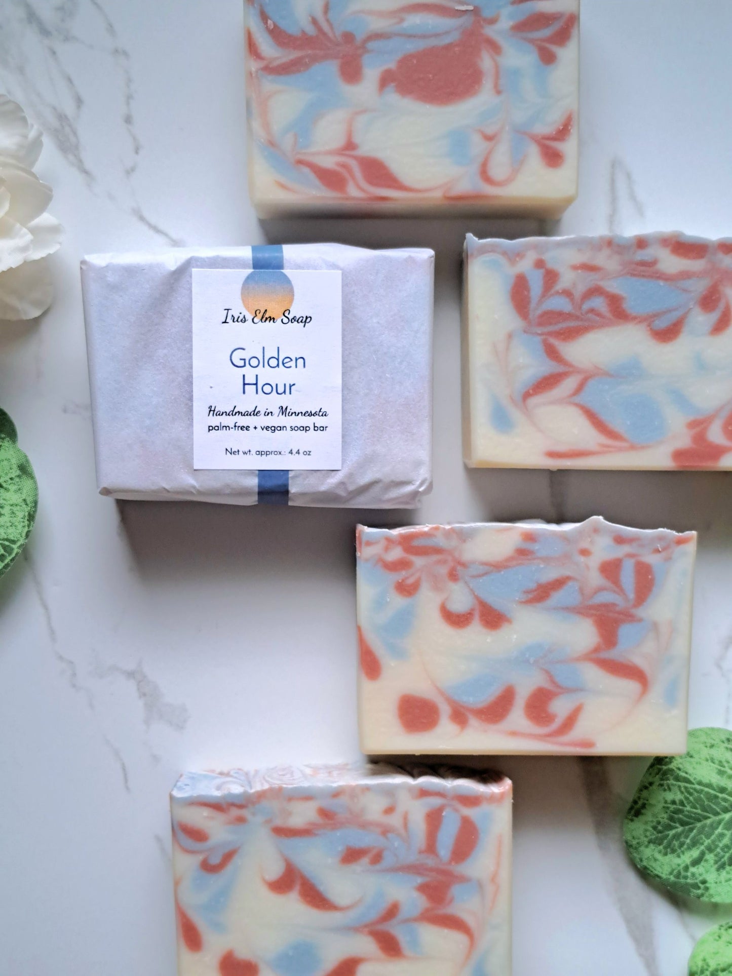 Five handmade soap bars with a blue, orange, and cream marble swirl pattern are arranged on a white marble surface. One soap bar is wrapped in white paper with a label that reads 'Jazzy Lane Soap Golden Hour Handmade in Minnesota palm oil + sugar soap bar'.