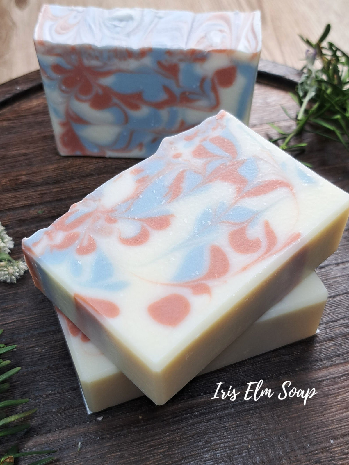 Three handmade marbled soap bars with cream, light blue, and dusty pink swirls, stacked on a dark wooden surface with a sprig of rosemary and a second soap bar in the background. The text 'Iris Elm Soap' is visible in the bottom right corner.