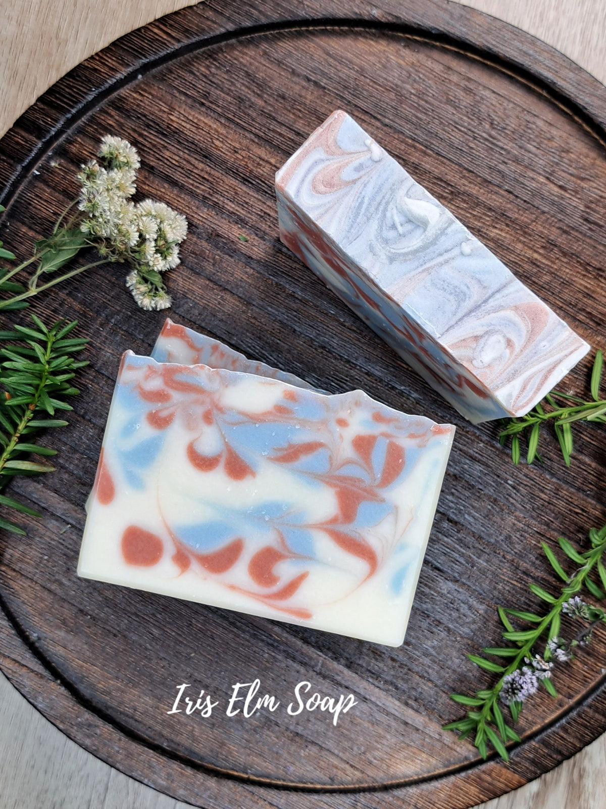 Two artisanal soap bars with a marbled swirl pattern of white, light blue, and terracotta orange, styled on a dark round wooden tray with green sprigs and small white flowers. The brand name 'Iris Elm Soap' is written in white script at the bottom.