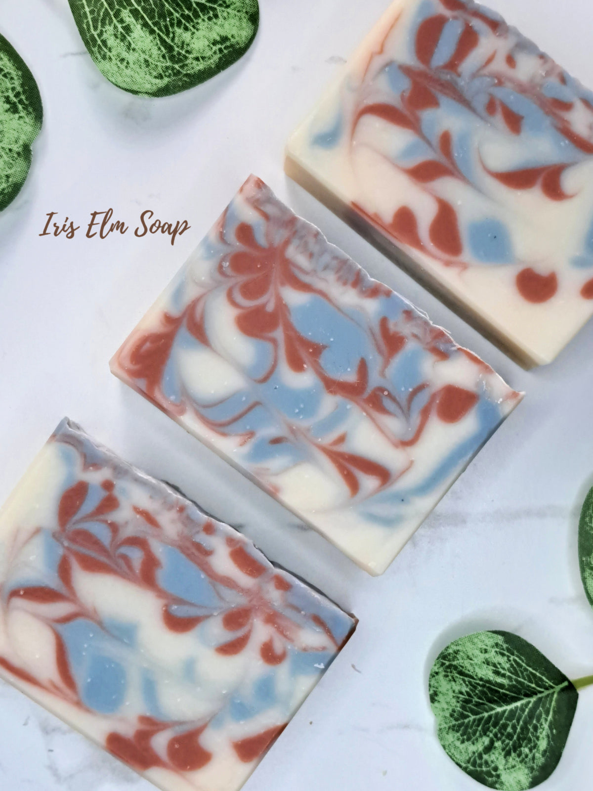 Three rectangular bars of swirled soap with cream, light blue, and terracotta red patterns, arranged diagonally on a white marble surface with green leaves. The text 'Iris Elm Soap' is visible on the left.