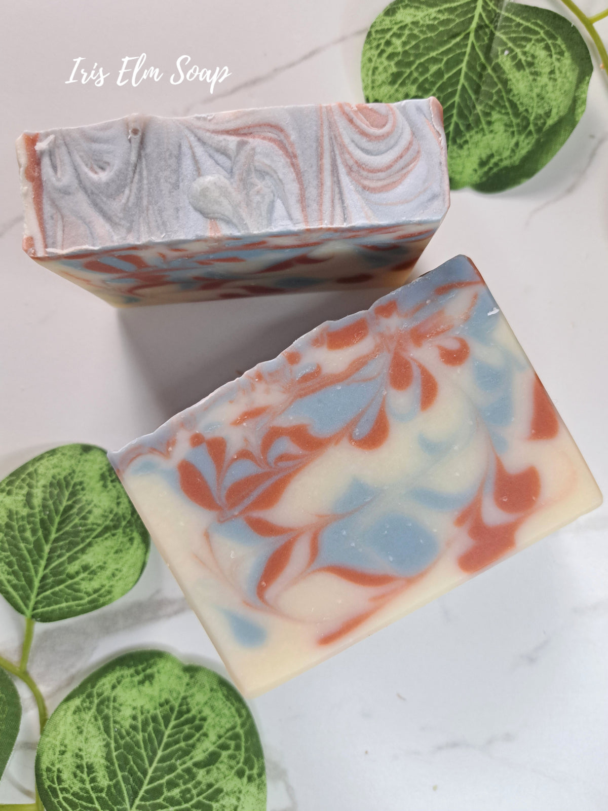 Two rectangular artisan soap bars with marbled swirls of cream, light blue, and coral-red are arranged on a white marble surface with green leaves. The brand name 'Iris Elm Soap' is written in white script in the upper left.