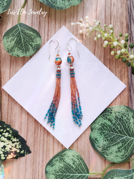 Beaded orange and teal ocean jade tassel earrings on a white napkin with green leaves around