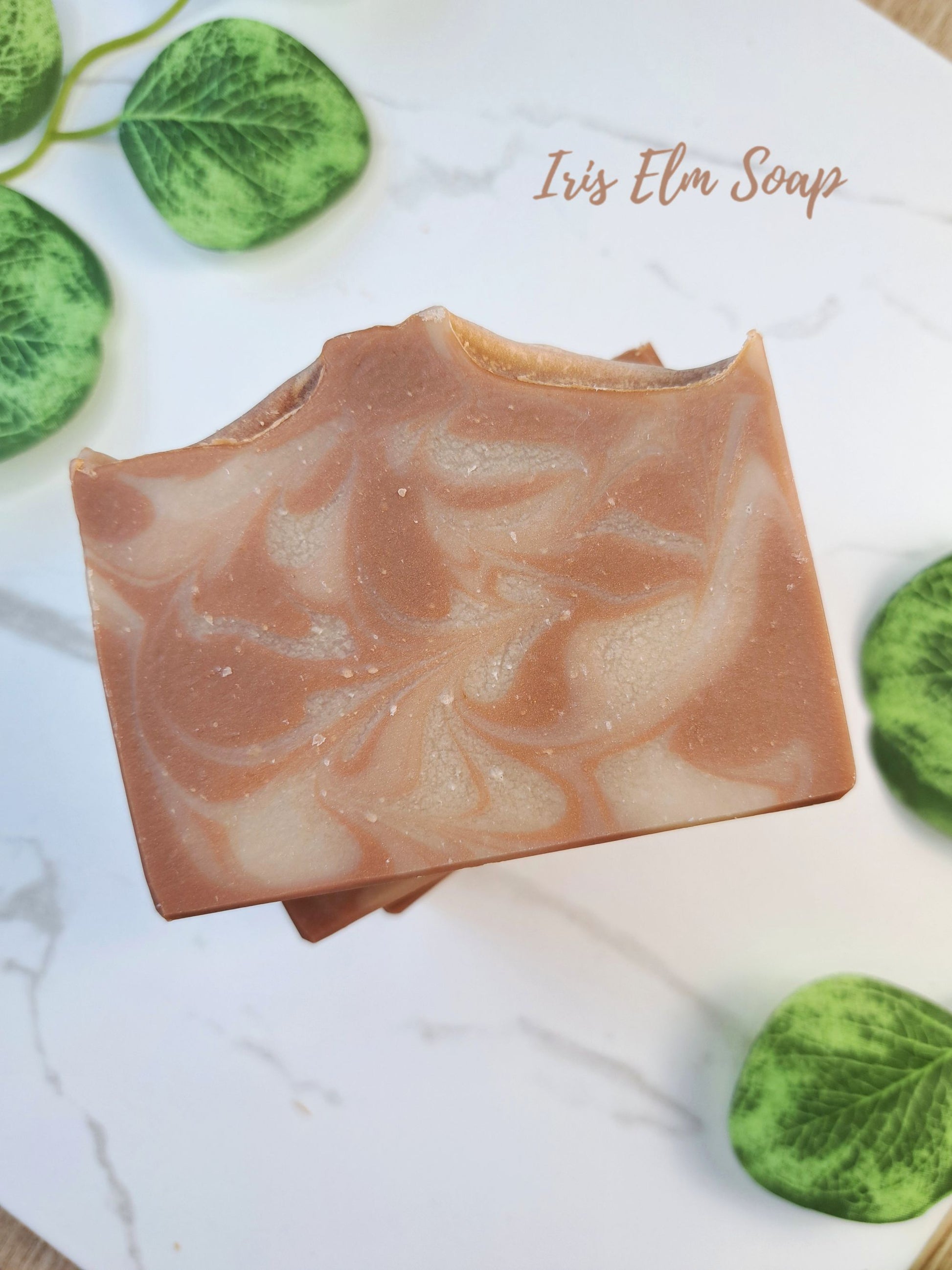 A bar of handcrafted oatmeal chai latte soap with a swirled light brown and cream pattern, displayed on a white marble surface with green decorative leaves. The text 'Iris Elm Soap' is written in the upper right.