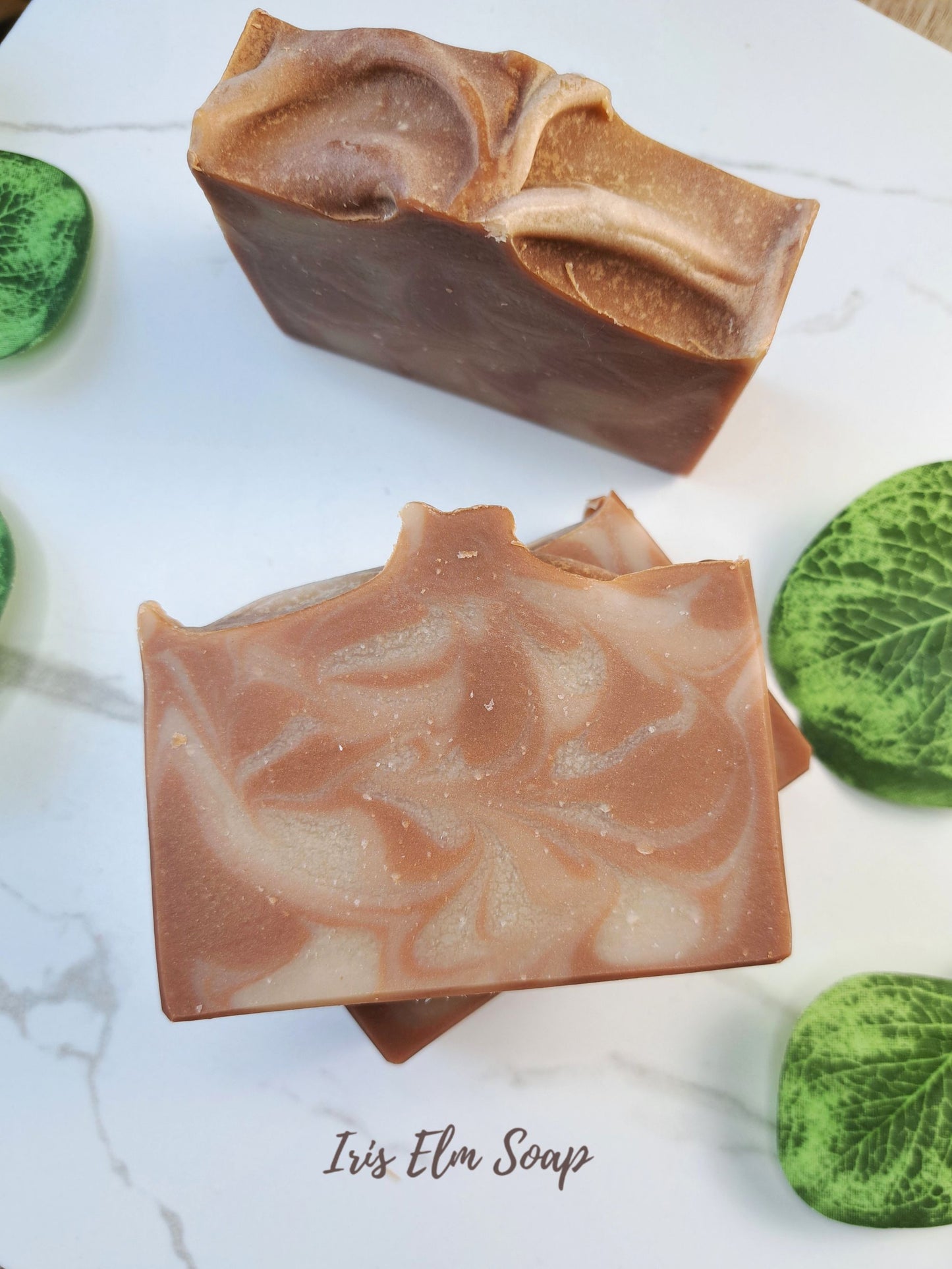 Two oatmeal chai latte artisanal soap bars with a brown and beige marbled swirl pattern, displayed on a white marble surface with green decorative stones. The text 'Iris Elm Soap' is visible at the bottom.
