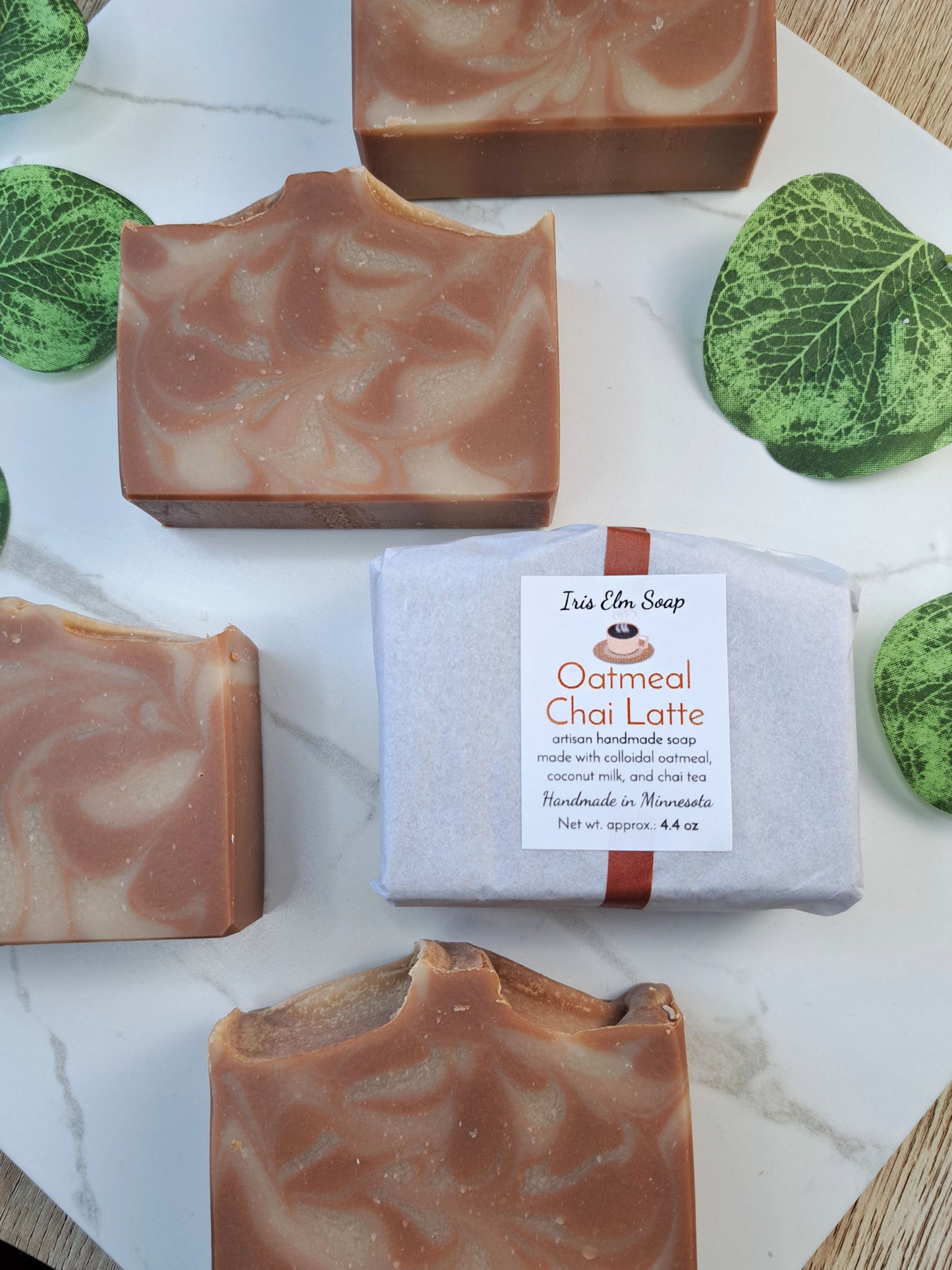Several rectangular bars of oatmeal chai latte soap are arranged on a white marble surface, with one bar wrapped in white paper and tied with a brown ribbon in the center. The soap has a warm tan and light brown marbled swirl.