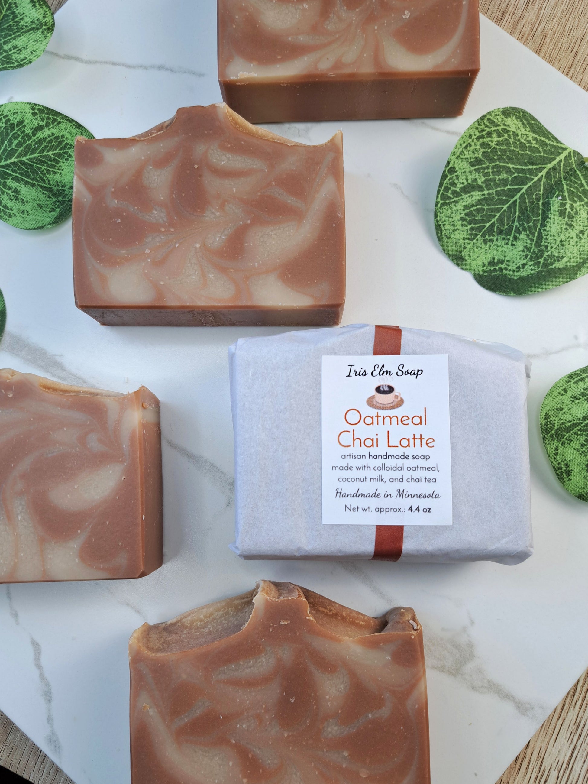 Several rectangular bars of oatmeal chai latte soap are arranged on a white marble surface, with one bar wrapped in white paper and tied with a brown ribbon in the center. The soap has a warm tan and light brown marbled swirl.