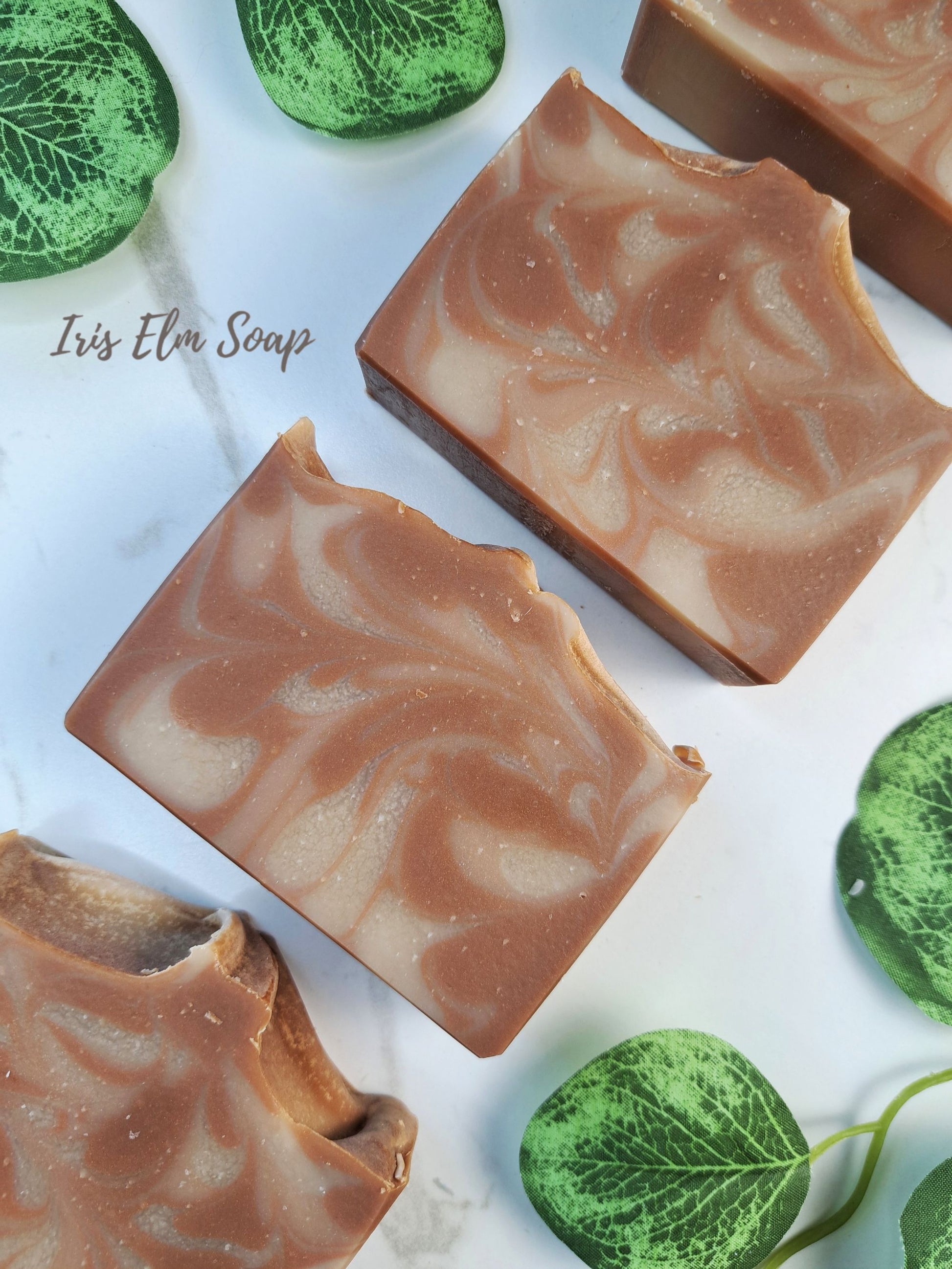 Four rectangular bars of artisanal oatmeal chai latte soap with a brown and cream marbled swirl pattern, arranged on a white marble surface with decorative green leaves. The text 'Iris Elm Soap' is visible on the left.