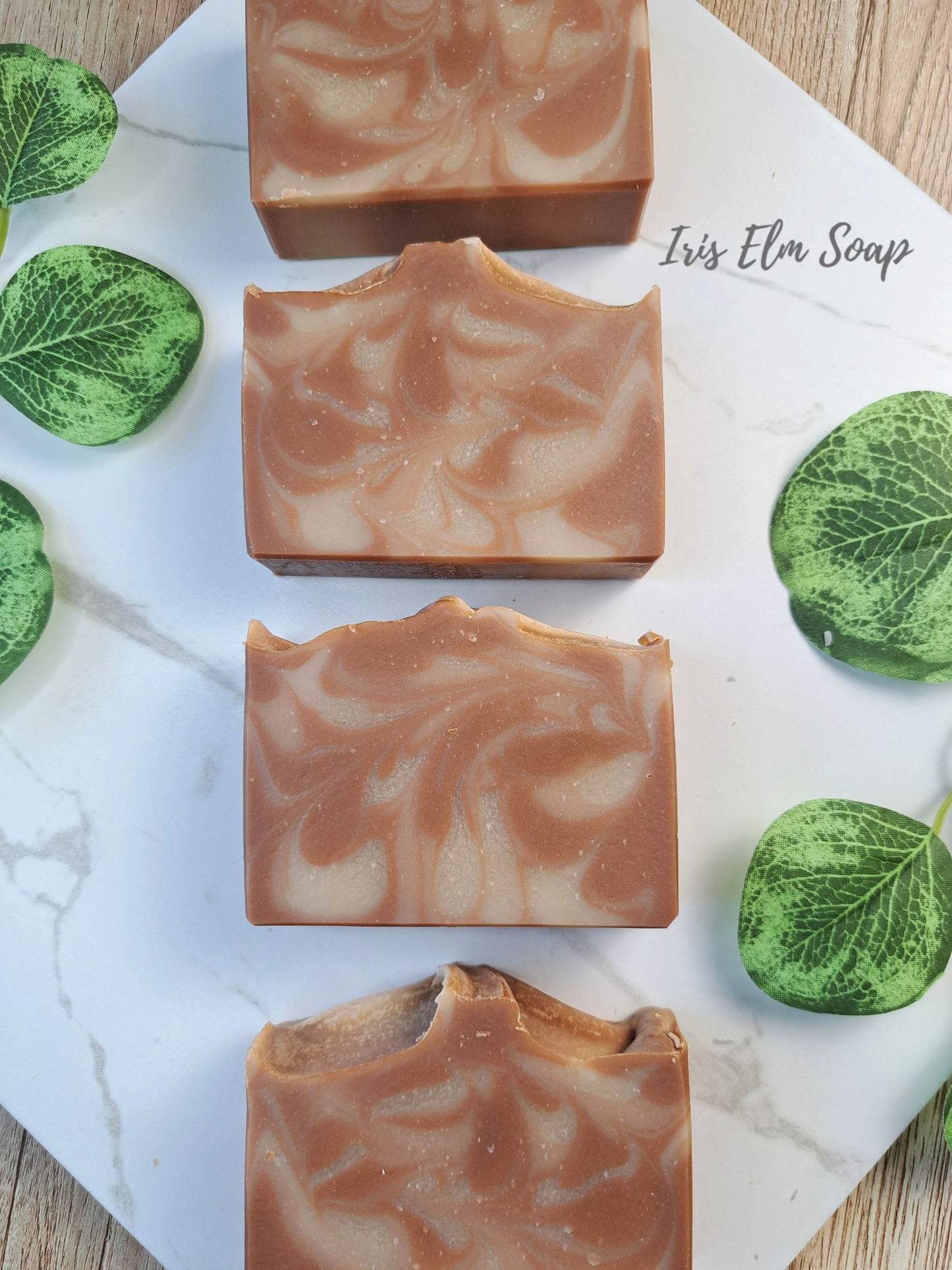 Four rectangular bars of swirled brown and cream oatmeal chai latte soap arranged vertically on a white marble board, with green decorative leaves around them and the text 'Iris Elm Soap' visible near the top right.