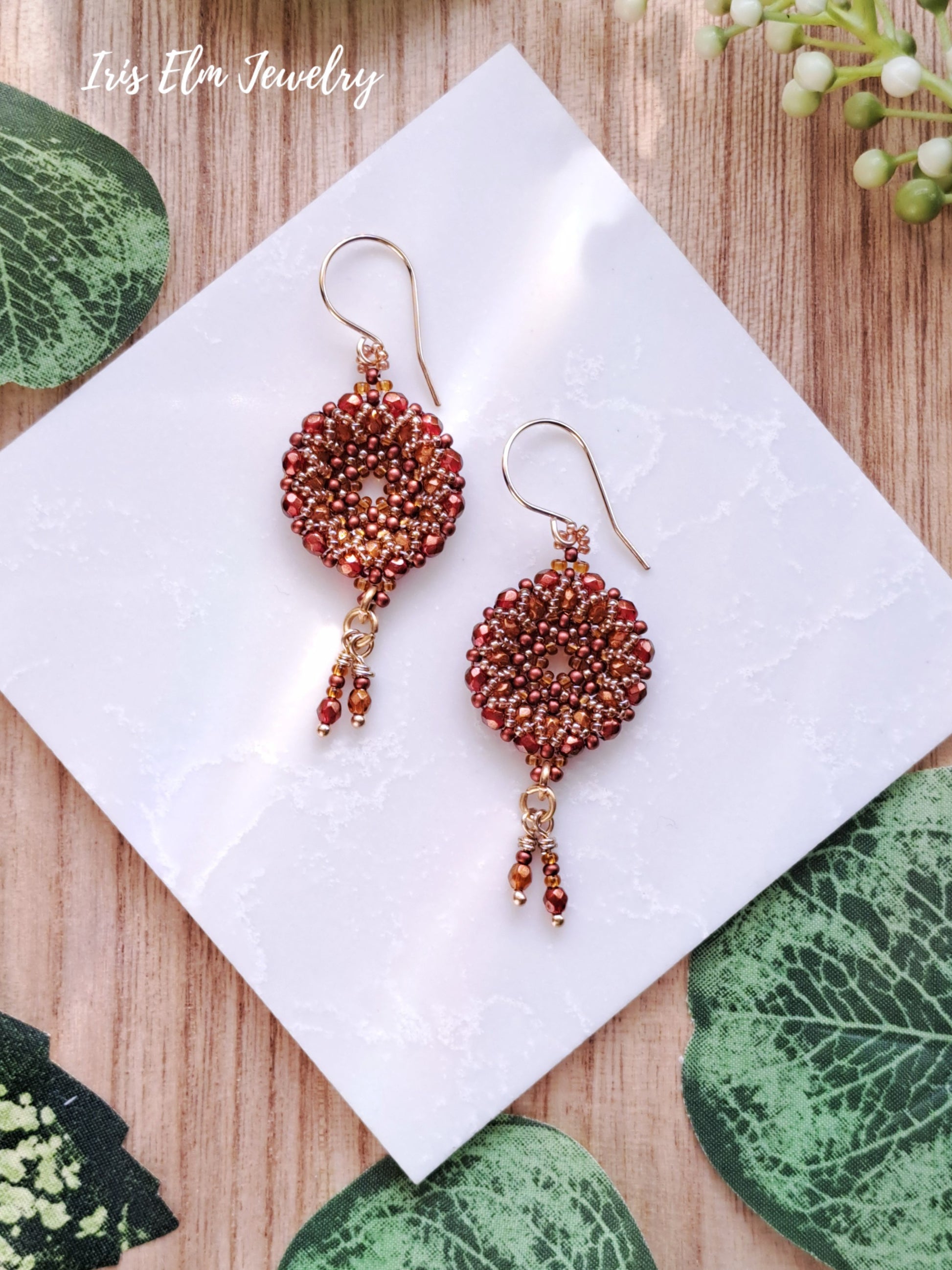 Pair of beaded red orange czech glass woven earrings on a white tile with green leaves around