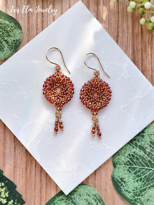 Pair of beaded red orange beadwork glass hoop wreath earrings on a white tile with green leaves in the background, featuring 'Iris Elm Jewelry'.