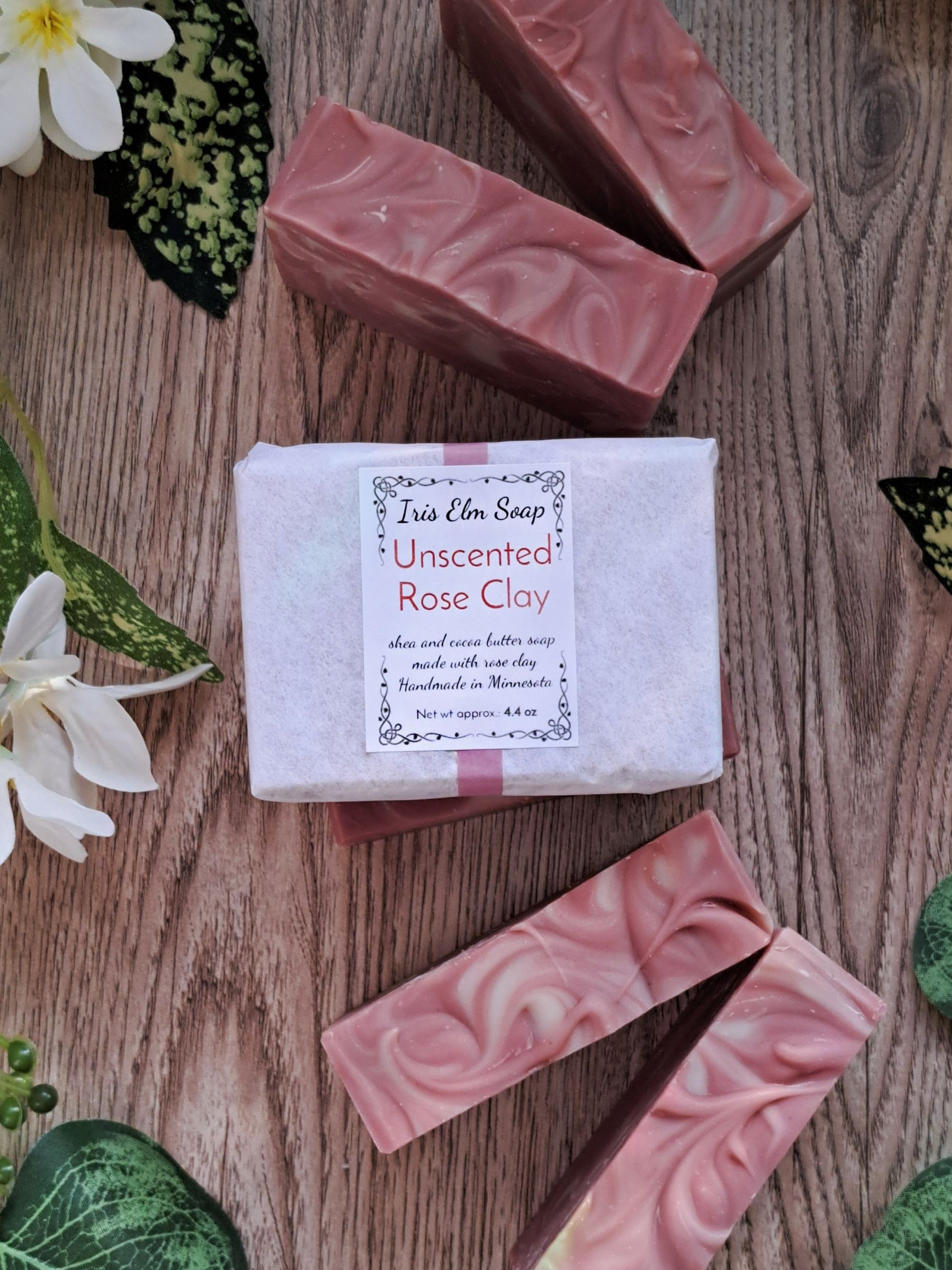 Unscented Rose Clay soap bar with a label on a wooden surface surrounded by flowers and leaves.