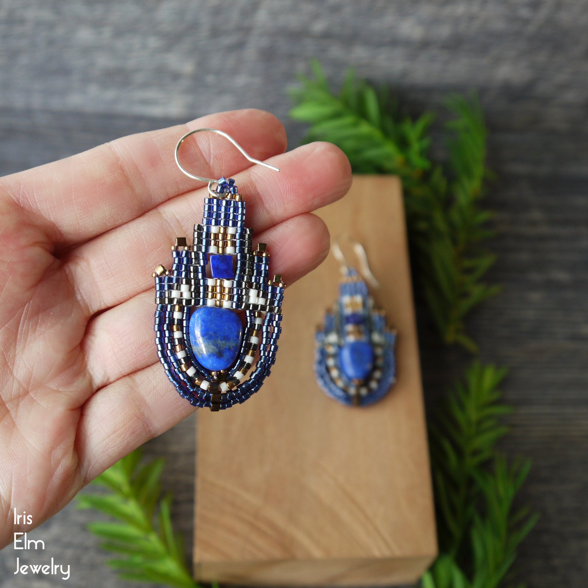 Blue lapis lazuli stone and seed bead beadwork teardrop earrings held in a hand with a blurred background