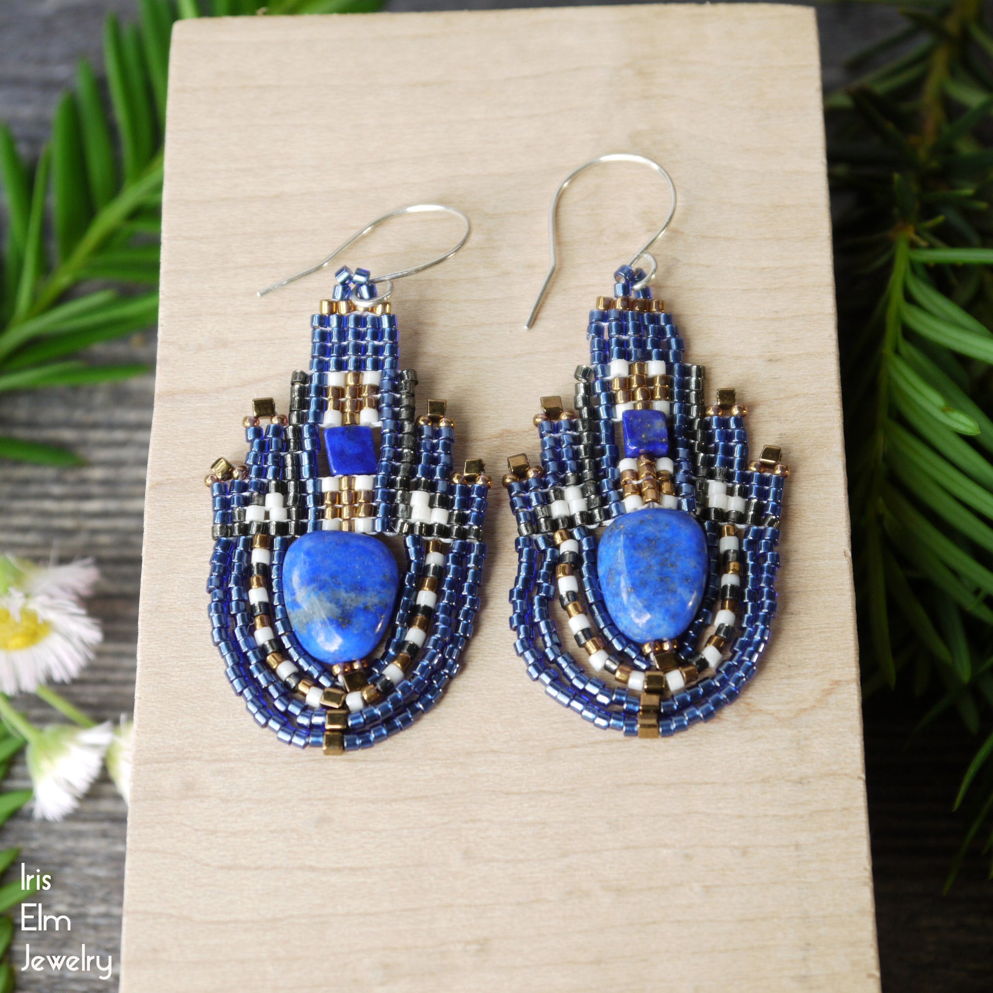 Blue lapis lazuli stone and seed bead teardrop beaded earrings on a wooden surface with greenery in the background