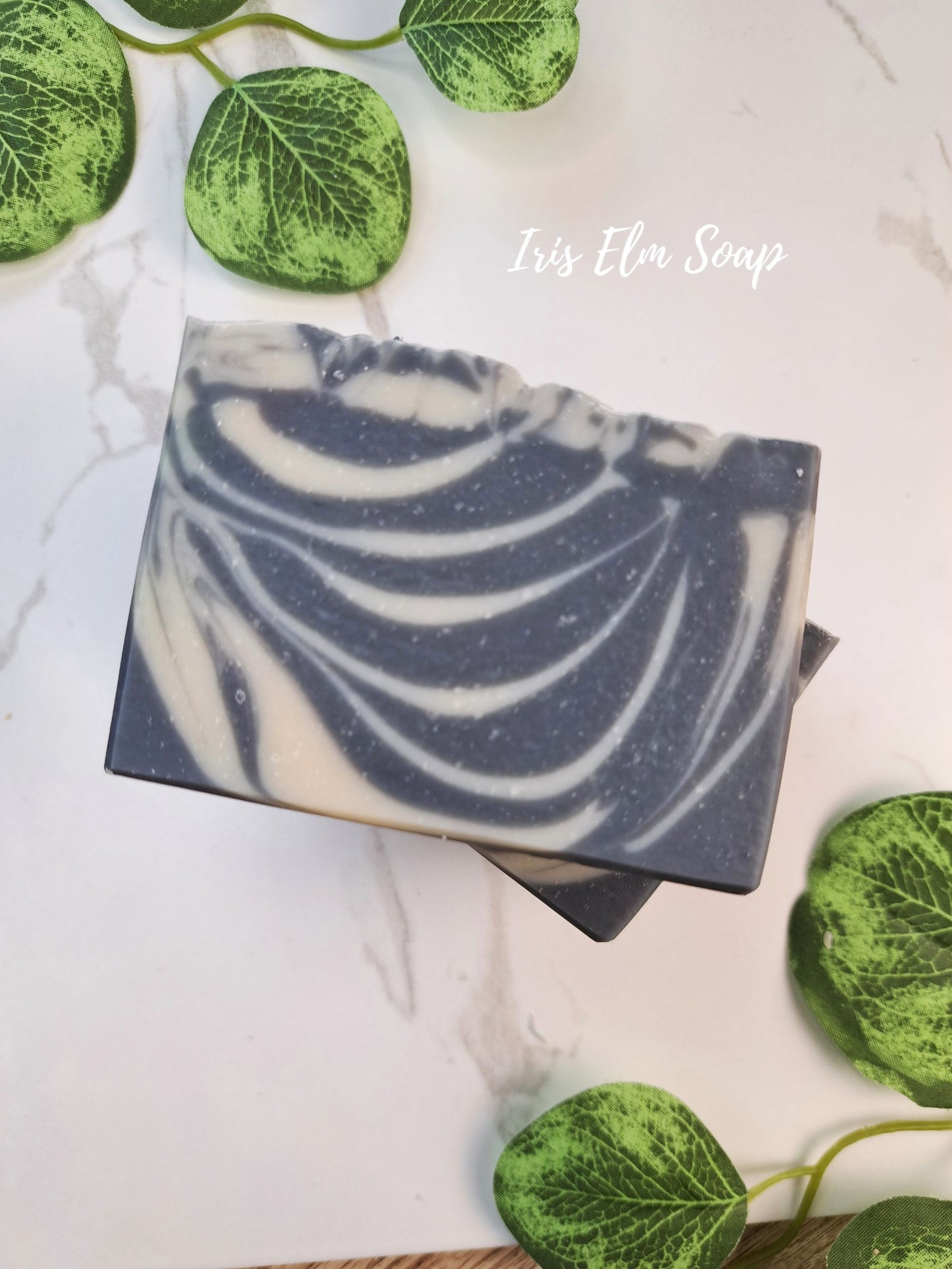 A rectangular bar of soothing sandalwood soap with a zebra stripe swirl pattern of dark grey and cream, styled on a white marble surface with green decorative leaves. The text 'Iris Elm Soap' is written in white script in the upper right.