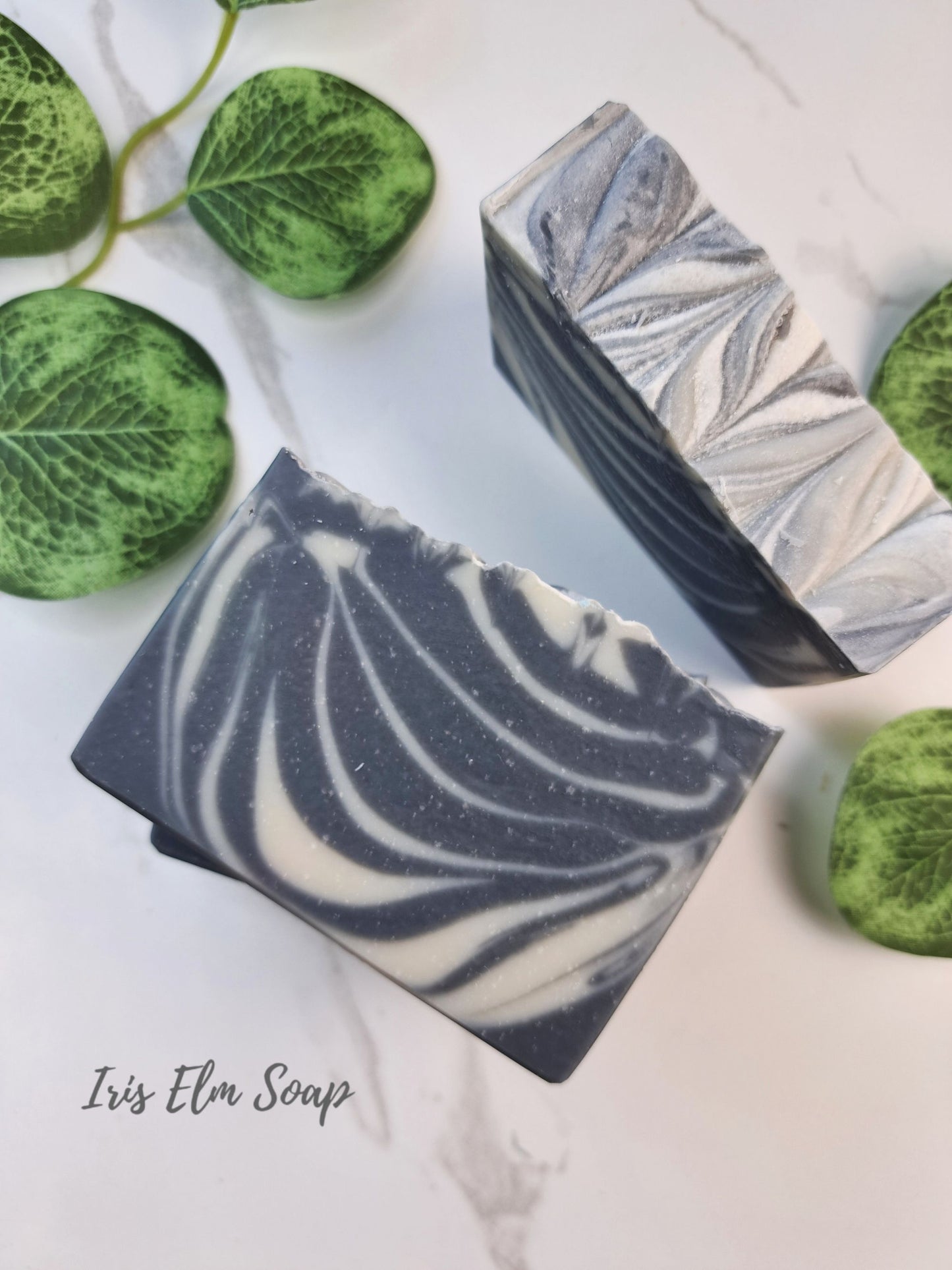 Two rectangular soothing sandalwood bars of Iris Elm Soap with a black and white marbled swirl pattern, styled on a white marble background with green decorative leaves.
