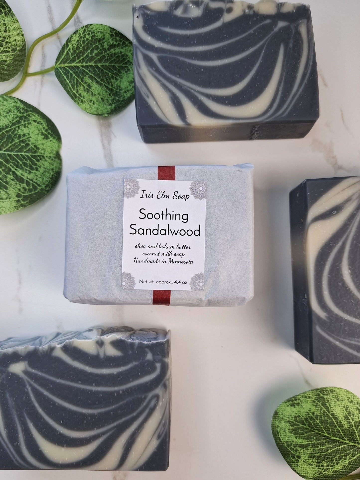 Three rectangular bars of Iris Elm Soap, including one wrapped in white paper with a 'Soothing Sandalwood' label, arranged on a white marble surface with green decorative leaves.