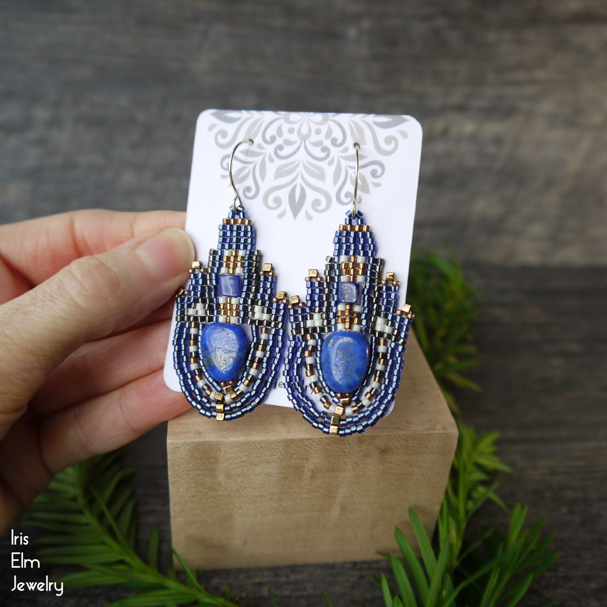Blue lapis lazuli beaded earrings on a wooden stand with a hand holding it, against a gray background.