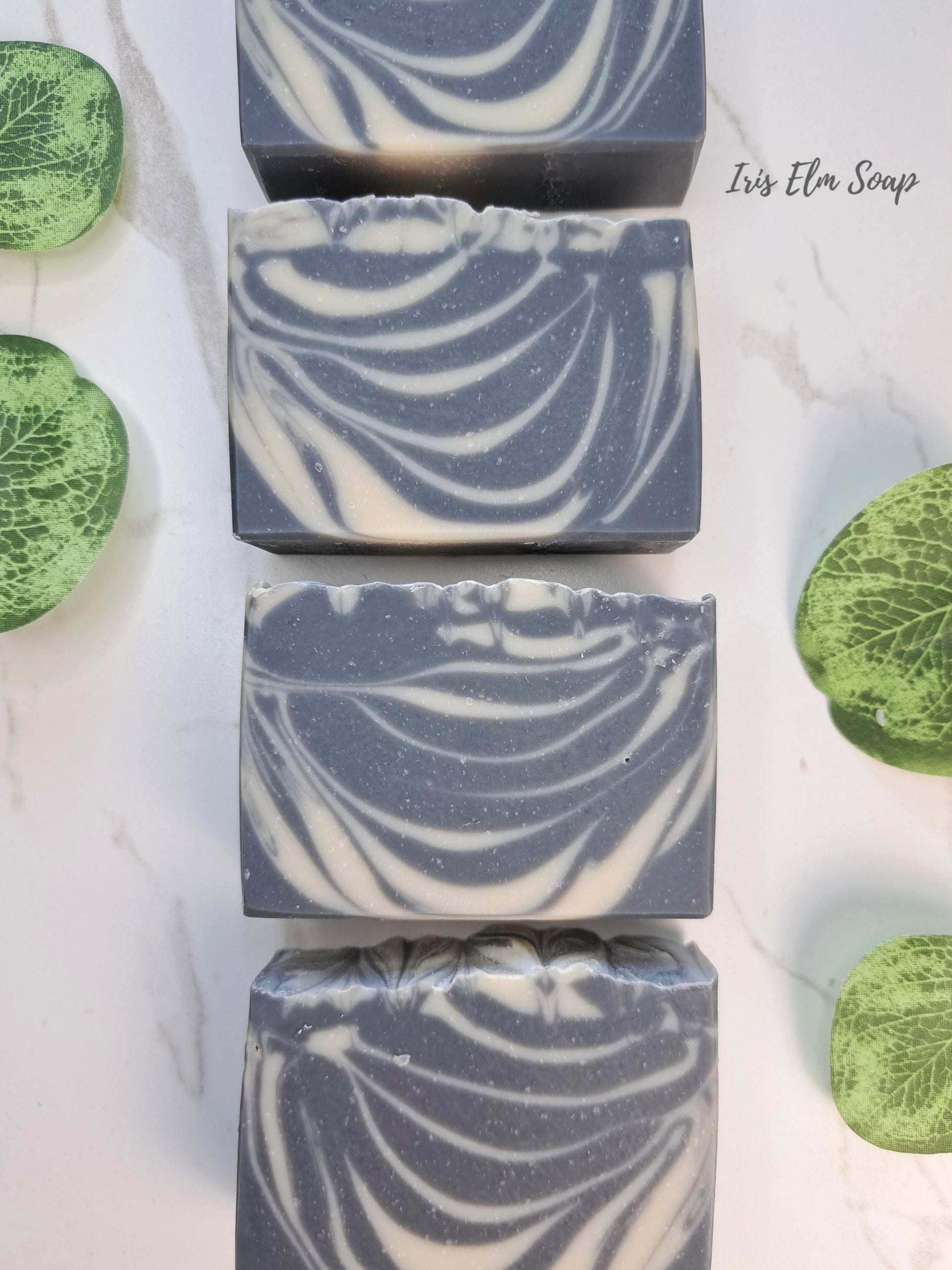 Four rectangular bars of soothing sandalwood artisanal soap with a blue-gray and white marbled swirl pattern, arranged vertically on a white marble surface with green leaf-shaped decorative props. The text 'Iris Elm Soap' is visible in the upper right.