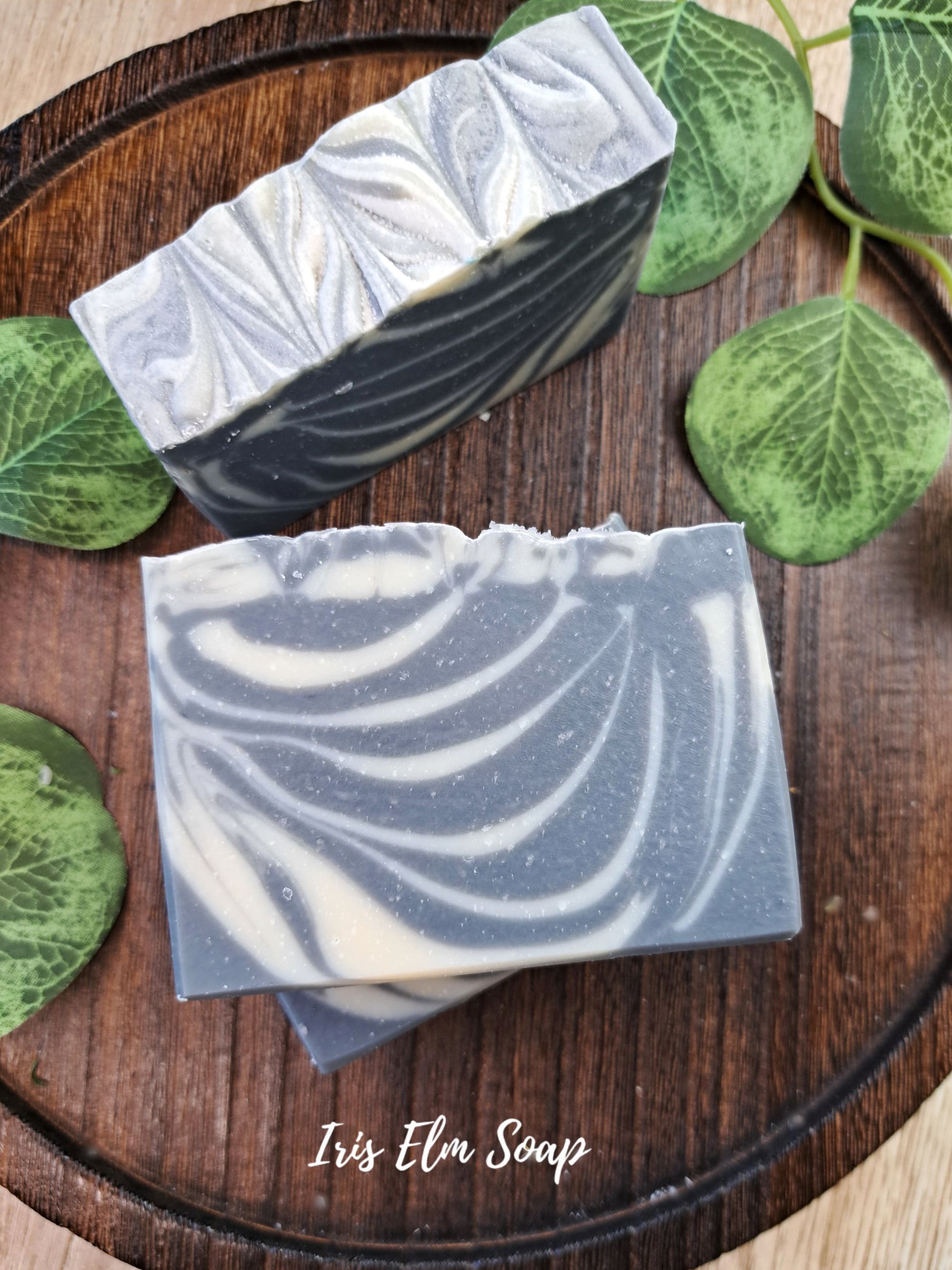Two artisanal handcrafted soothing sandalwood soap bars with a gray and white marbled swirl pattern, displayed on a dark wooden tray with green leaves. The brand name 'Iris Elm Soap' is visible as a text overlay at the bottom.