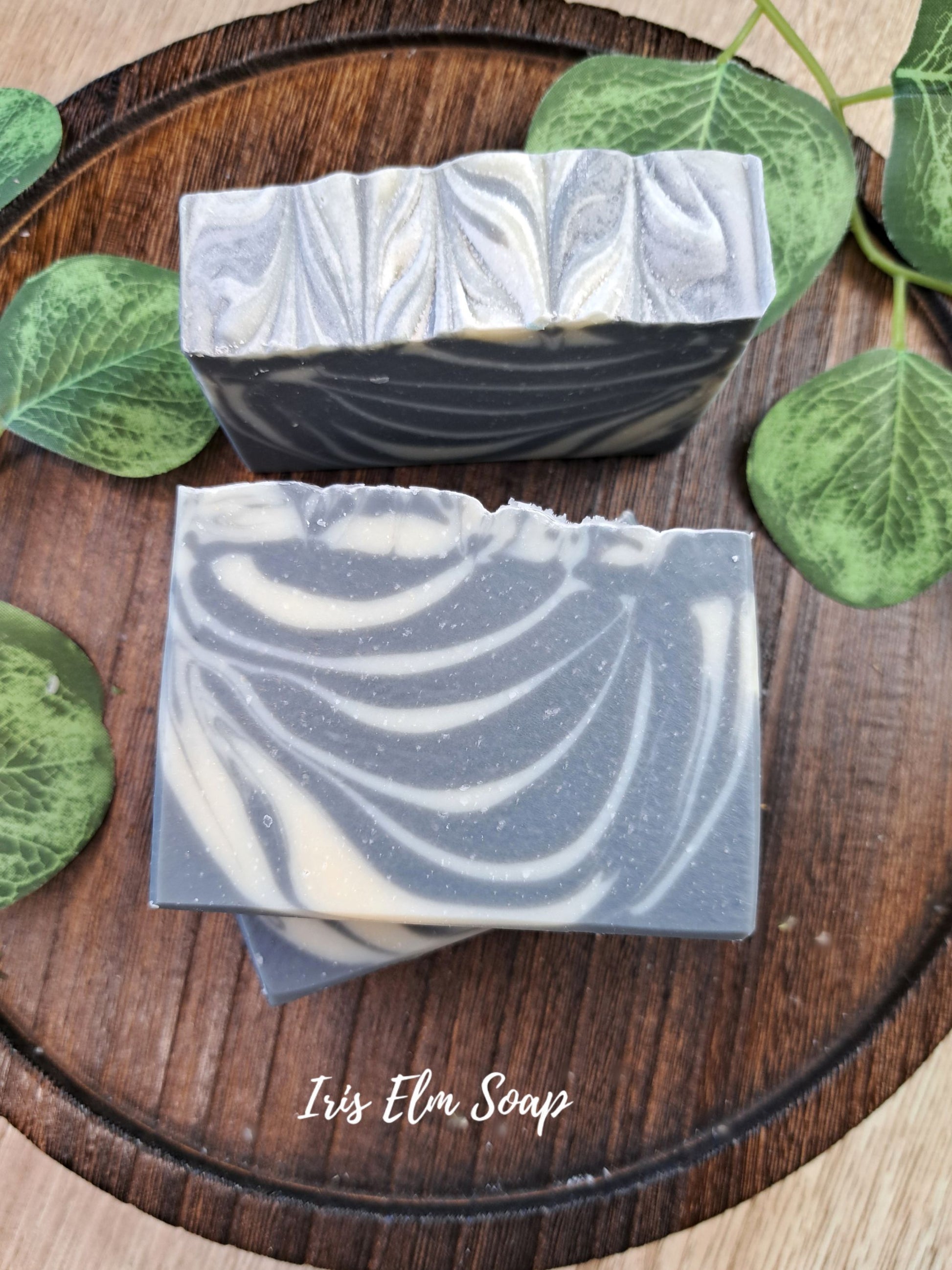 Two rectangular artisan soothing sandalwood soap bars with a gray and white marbled swirl pattern, stacked on a round dark wooden tray and surrounded by green leaves. The brand name 'Iris Elm Soap' is written in white script at the bottom.