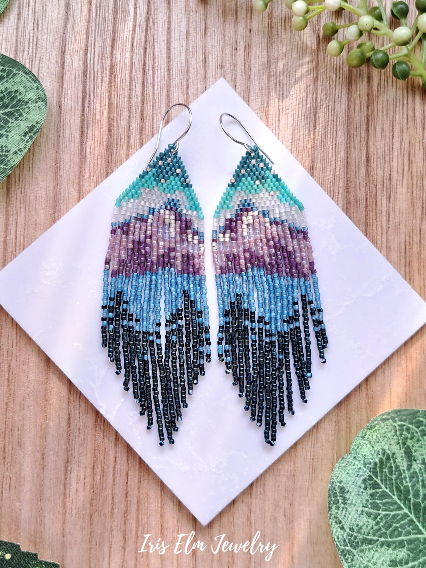 Beadwoven mountain forest lake seed bead fringe earrings surrounded by greenery with Iris Elm Jewelry branding