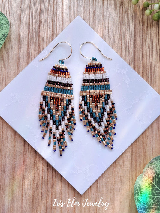 Beaded orange turquoise blue purple gold seed bead fringe boho earrings on a white square with 'Iris Elm Jewelry' branding, on a wooden surface.