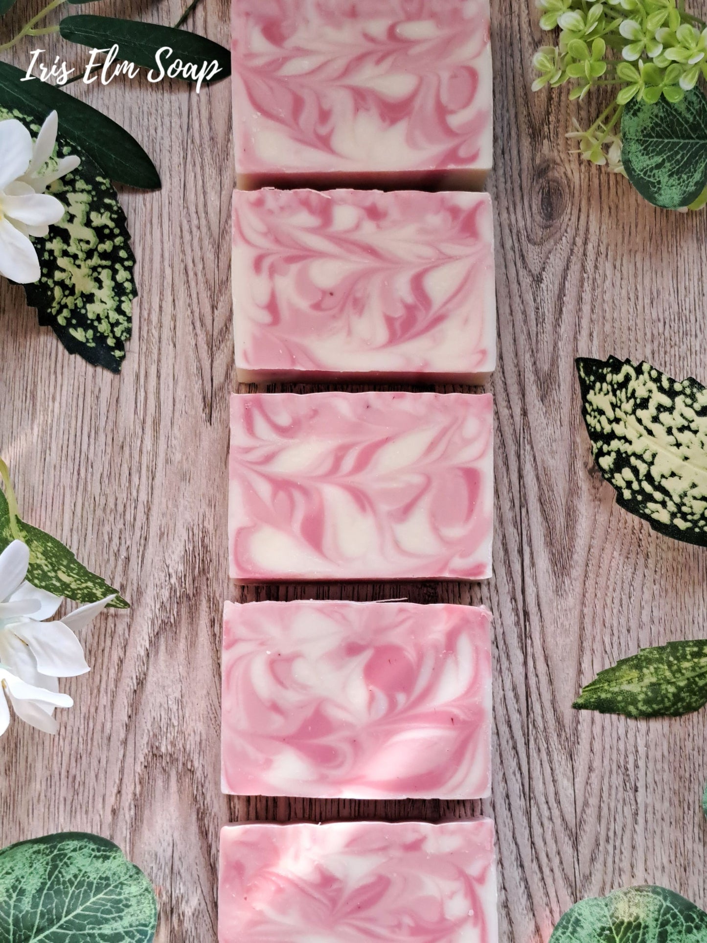 Pink and white swirled citrus love spell handmade soap bars on a wooden surface with green leaves and flowers.
