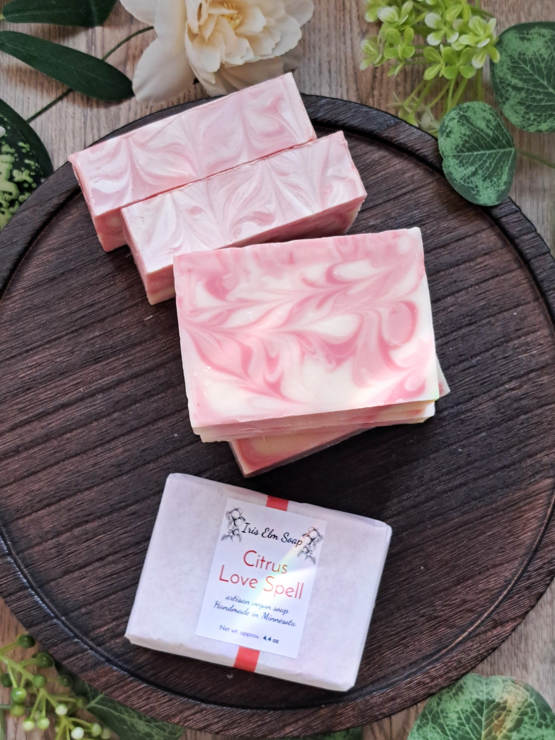 Three pink marbled citrus love spell handmade soap bars on a wooden board with greenery in the background