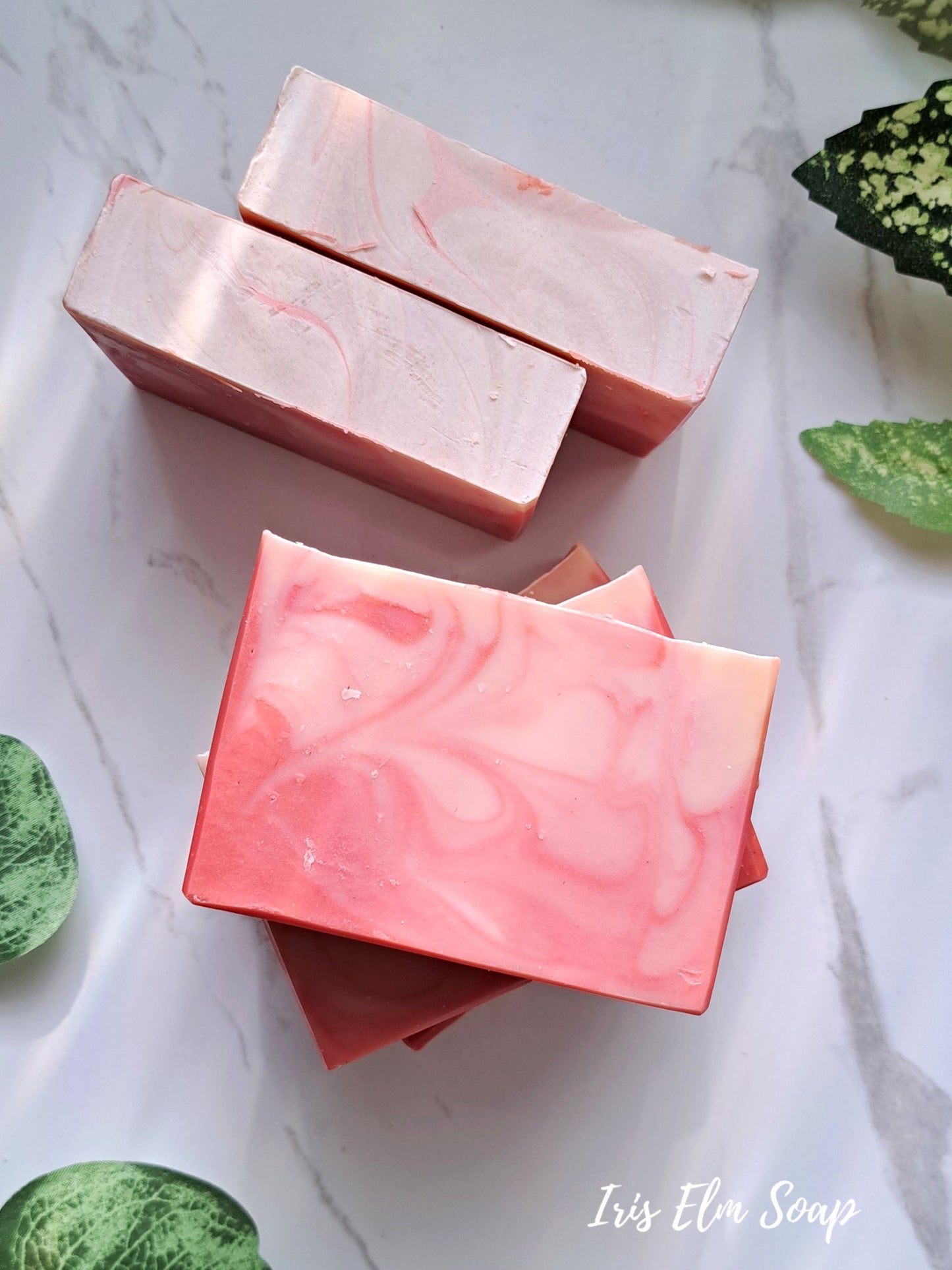 Three pink peach bellini handmade marbled soap bars on a marble surface with green leaves.