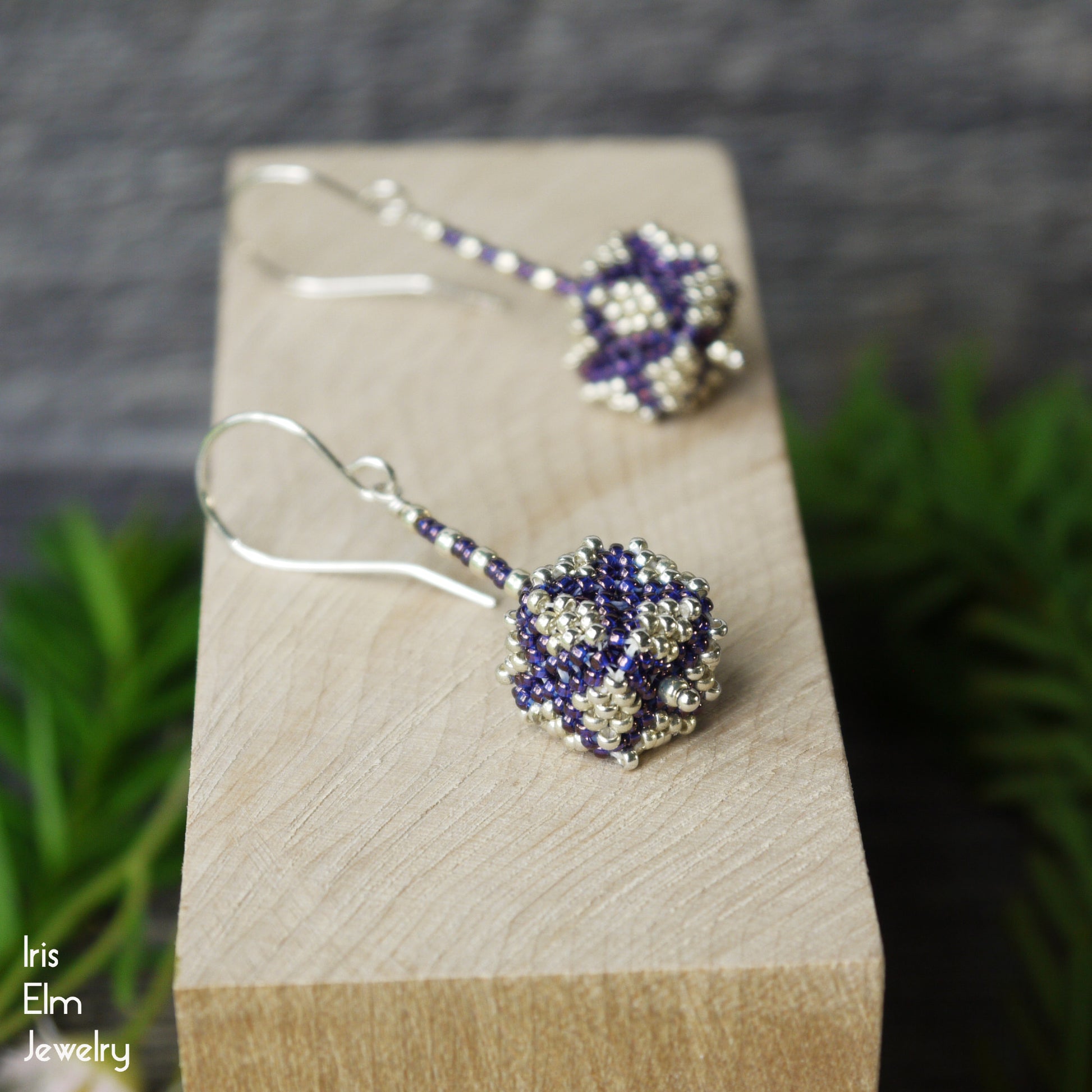 Beaded glass purple and silver seed bead cube shaped earrings on a wooden block with a blurred green plant background