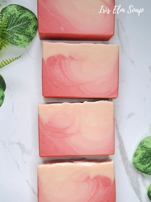Four rectangular bars of handcrafted peach bellini soap with pink and peach marbled swirls, arranged vertically on a white marble surface with green decorative leaves.