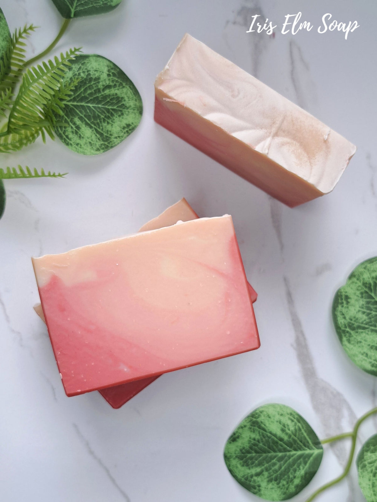 Two rectangular bars of peach bellini handcrafted soap with pink, peach, and cream marbled swirls, styled on a white marble surface with green decorative leaves. The brand name 'Iris Elm Soap' is written in white script in the top right corner.