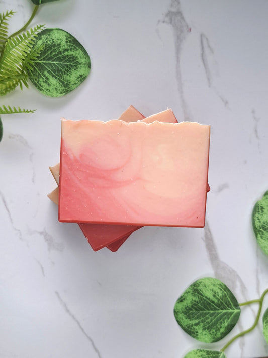 A stack of rectangular peach bellini handmade soap bars in soft coral, pink, and peach tones, arranged on a light marble surface with decorative green leaves around the edges.