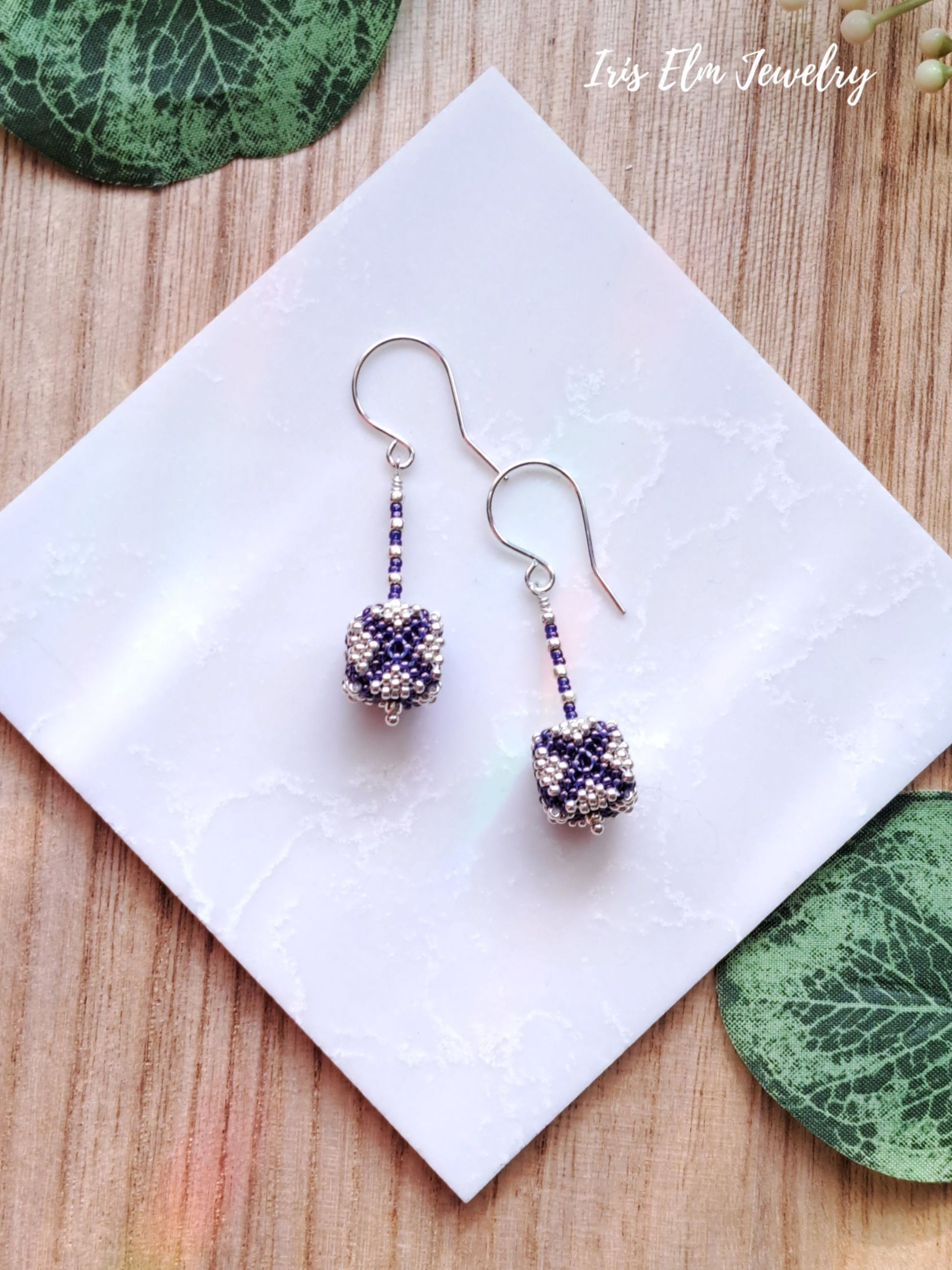 Pair of beaded purple and silver seed bead cube earrings on a white napkin with 'Iris Elm Jewelry' branding.