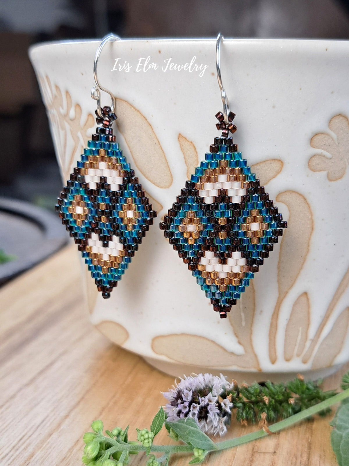 A pair of handmade beaded dangle earrings with a geometric diamond pattern in teal, brown, and cream, hanging from silver fishhook wires. They are displayed in front of a white ceramic cup on a wooden surface.