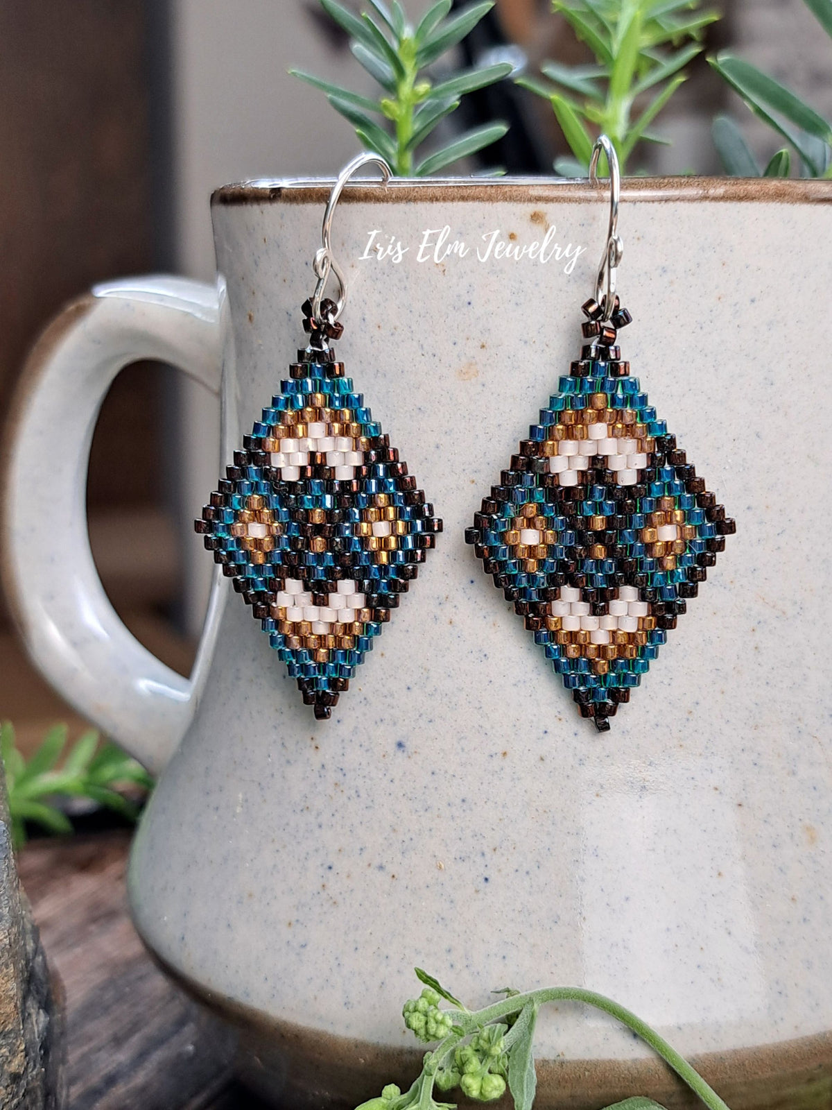 A pair of handmade beaded dangle earrings with a geometric diamond pattern in teal, brown, cream, hanging from silver hooks in front of a speckled ceramic mug.