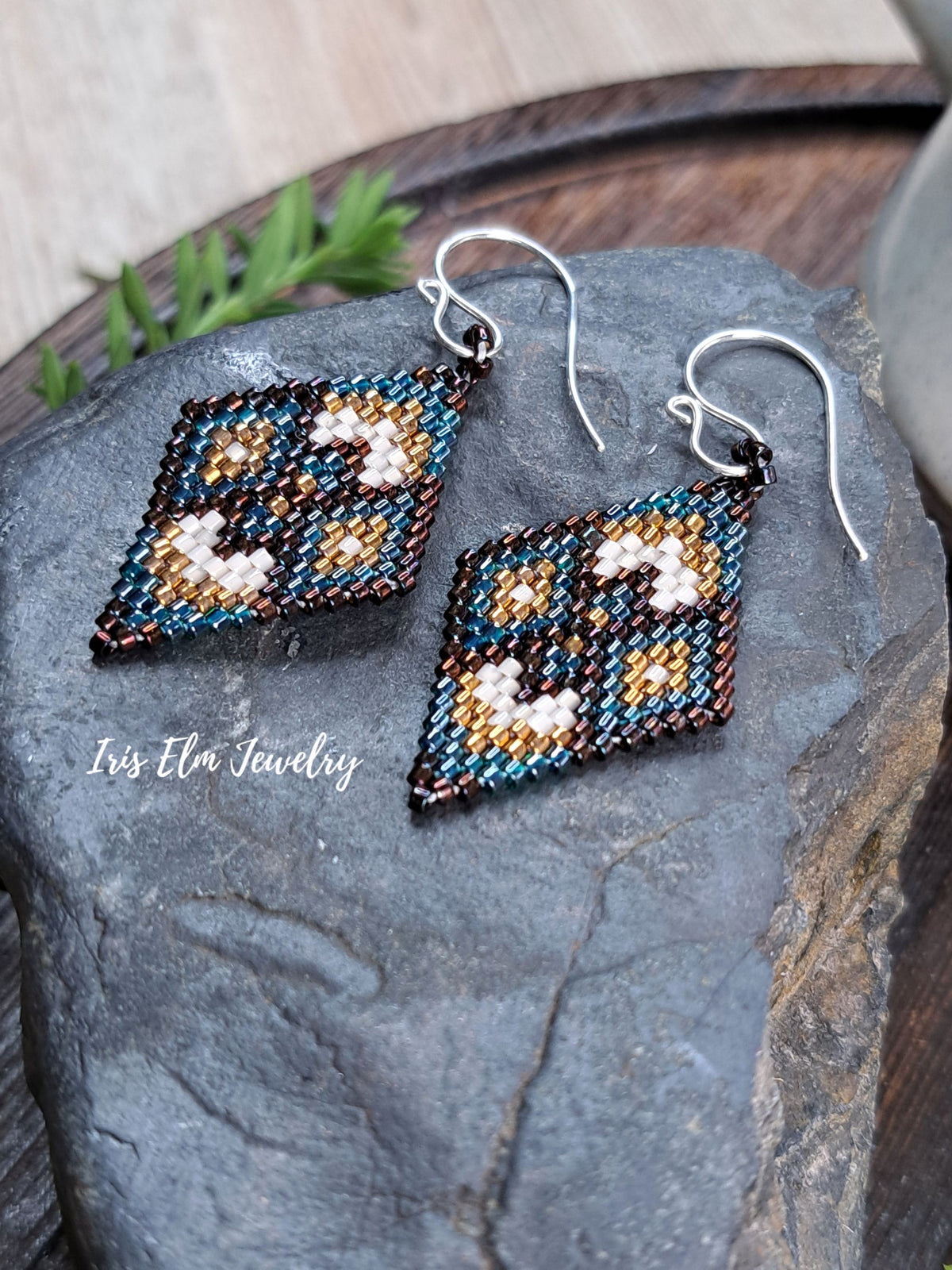 A pair of handmade beaded earrings with a geometric pattern in green, brown, gold, and cream, suspended from silver fishhook wires and displayed on a dark gray stone.