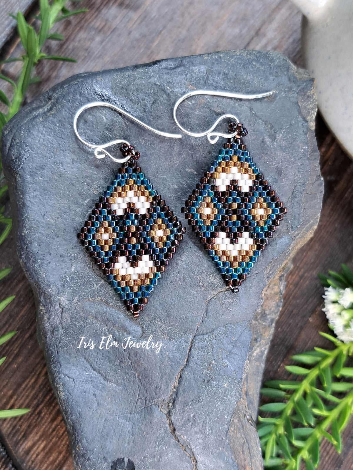 A pair of handmade beaded dangle earrings with a geometric pattern in teal, brown, cream, hanging from silver fishhook wires. They are displayed on a dark gray slate stone with green foliage and a wooden surface in the background.